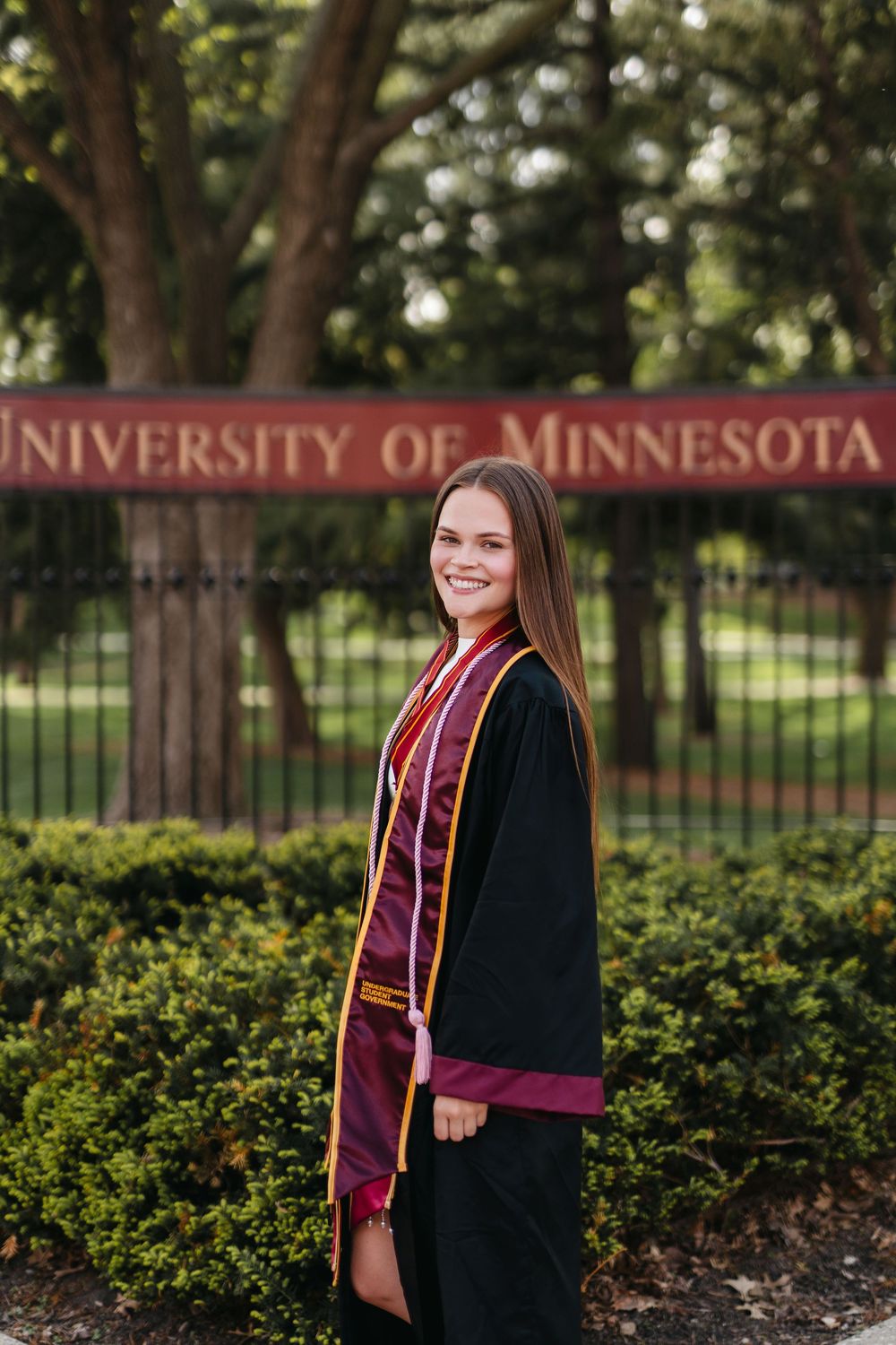 Graduate in cap and gown stands in front of University of Minnesota entrance sign surrounded by greenery.