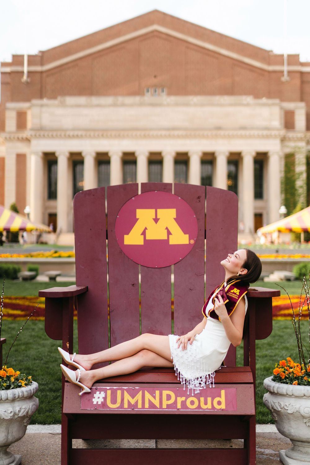 Graduate poses playfully on large maroon University of Minnesota chair with iconic Northrop Auditorium in background.