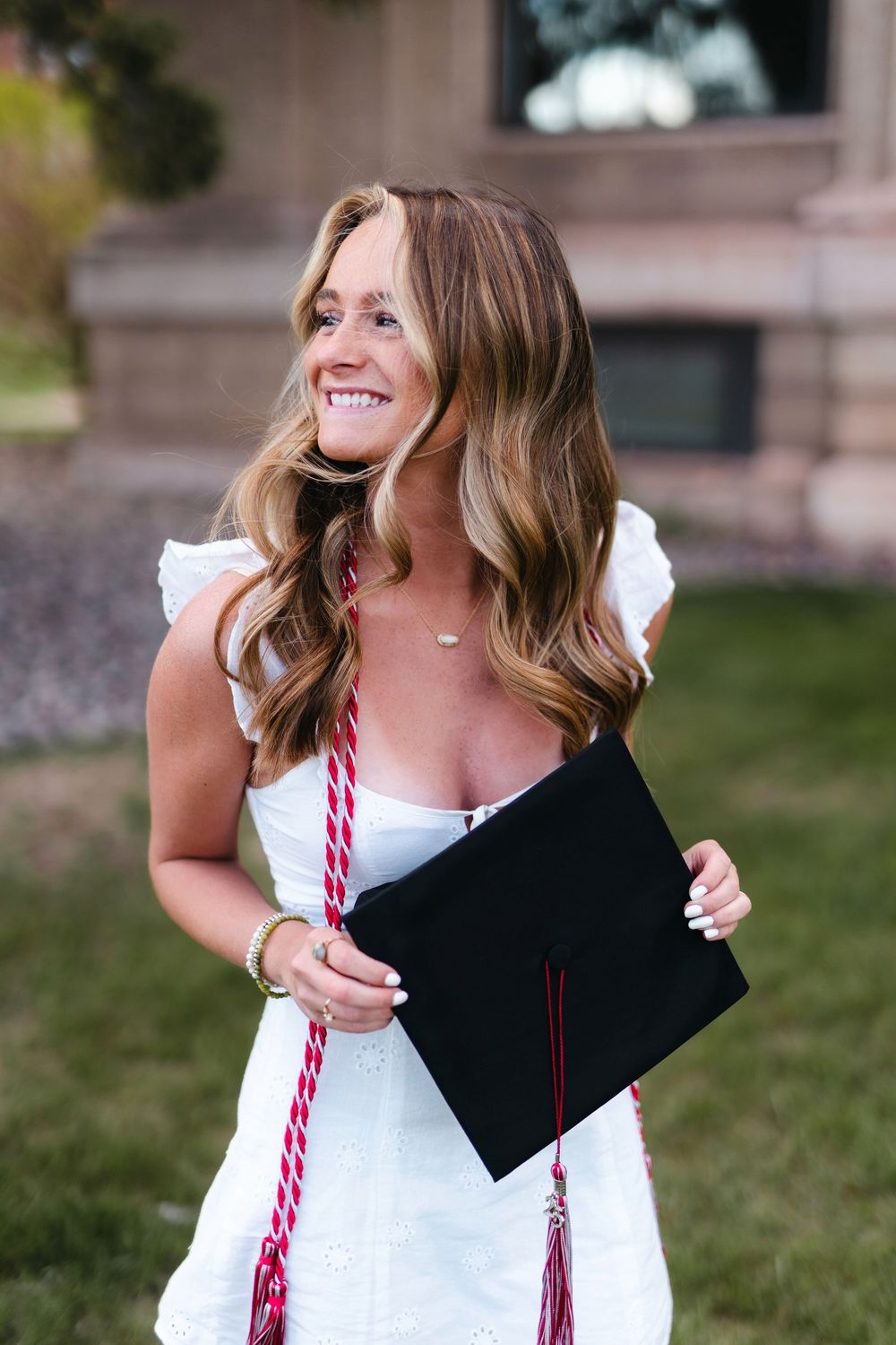 A graduate in a white dress holds a black diploma folder while posing outdoors for graduation photos.