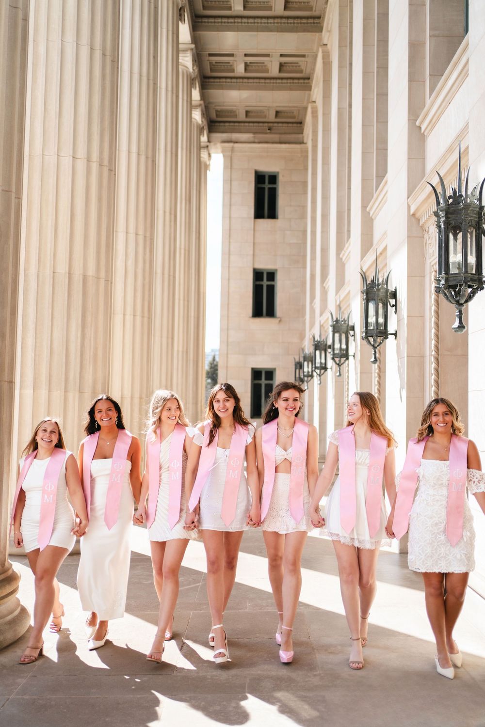 Group of women in pink dresses and white shoes walking along a columned corridor in elegant architectural setting.