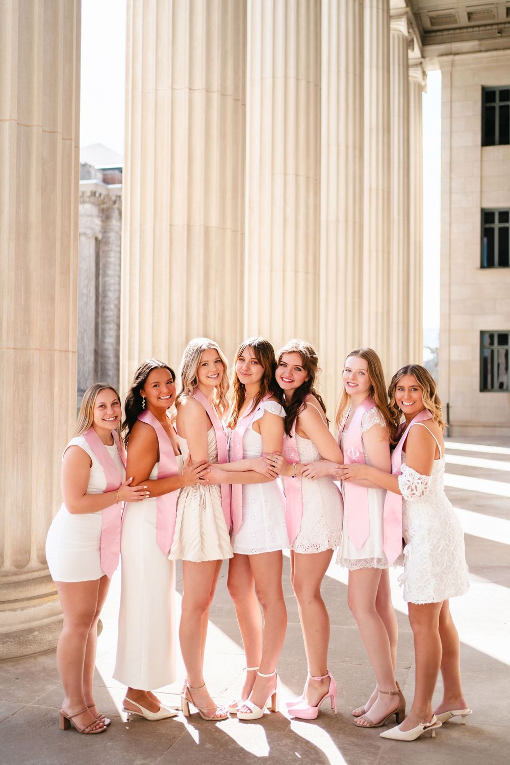 Group wearing pink dresses poses together in an elegant columned building interior.