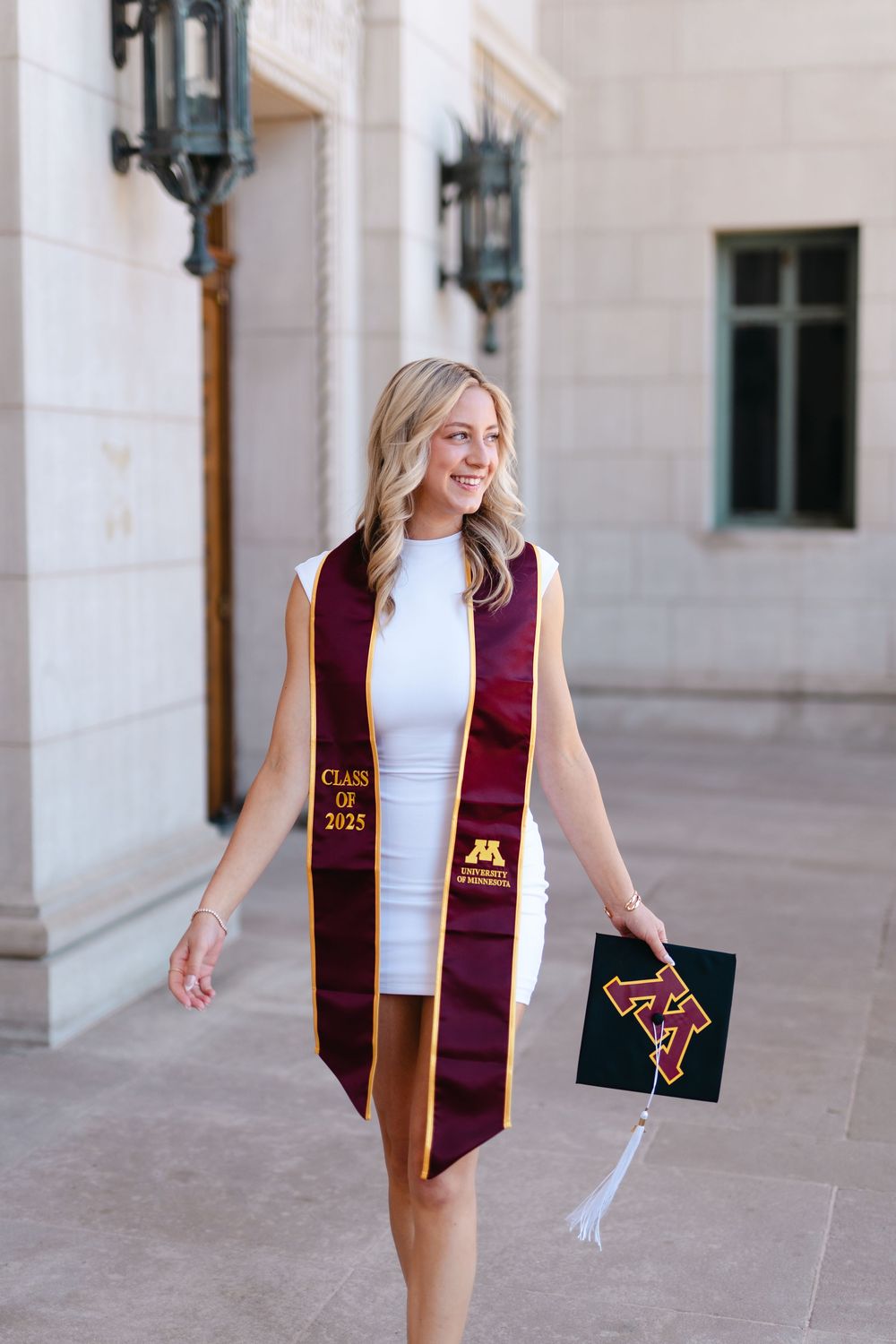 Graduate in white dress and burgundy stole walks through columned building during graduation photo session.