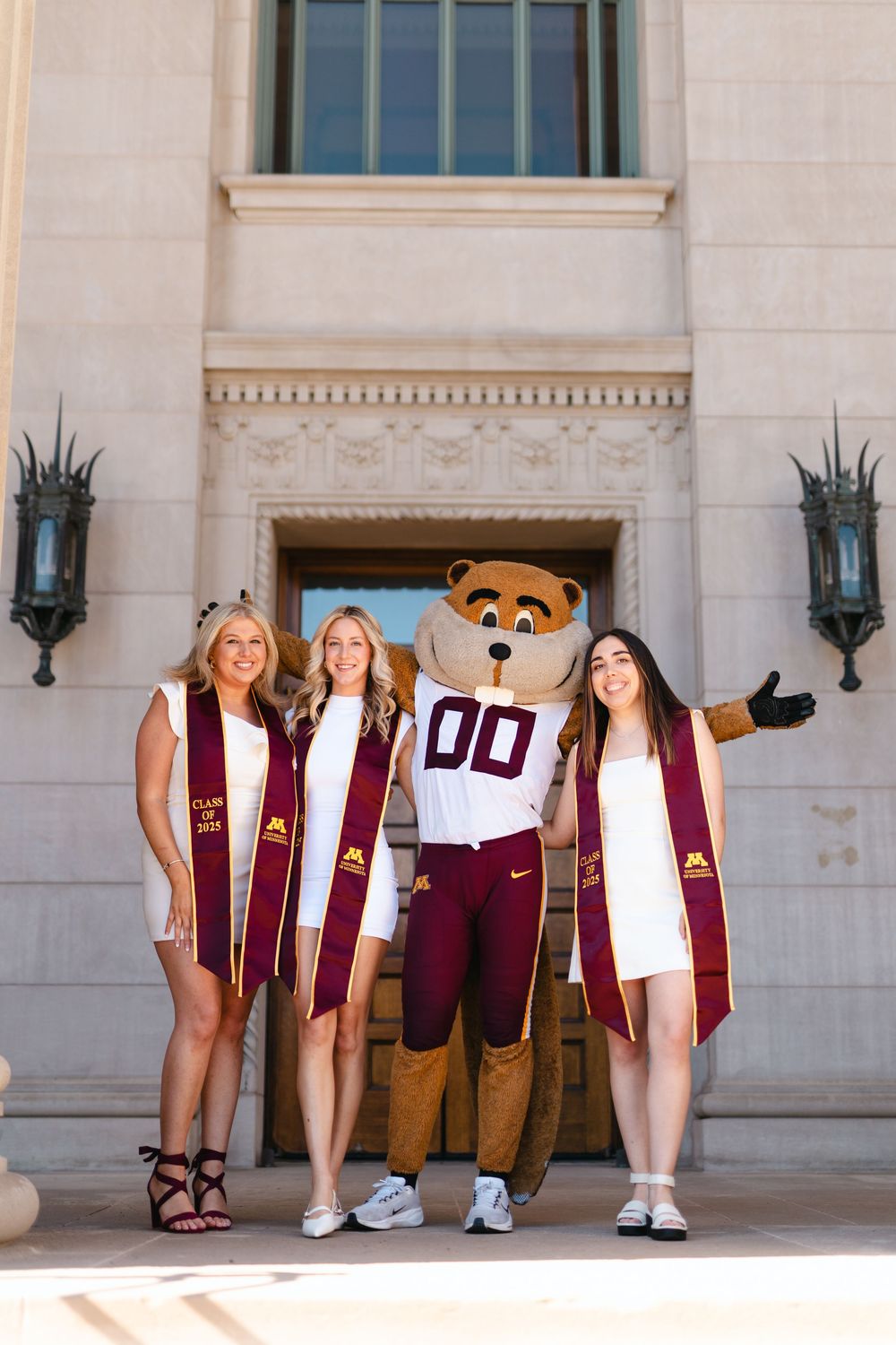 A creative graduation photo showing students and mascot lying on the ground with their feet against a university building wall.