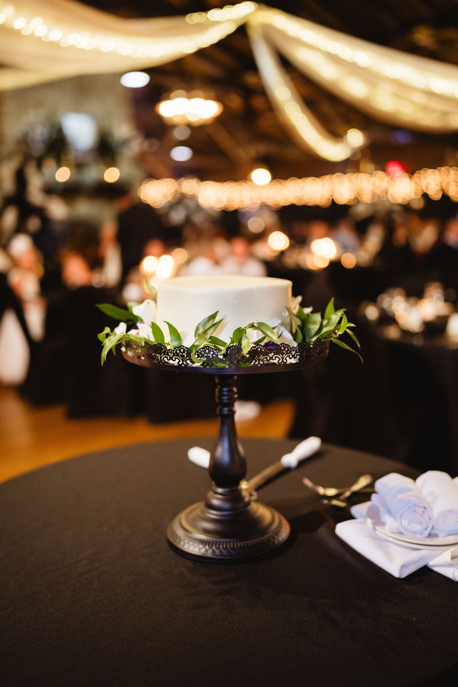 A white wedding cake displayed on an ornate black pedestal stand with greenery accents under romantic string lights.