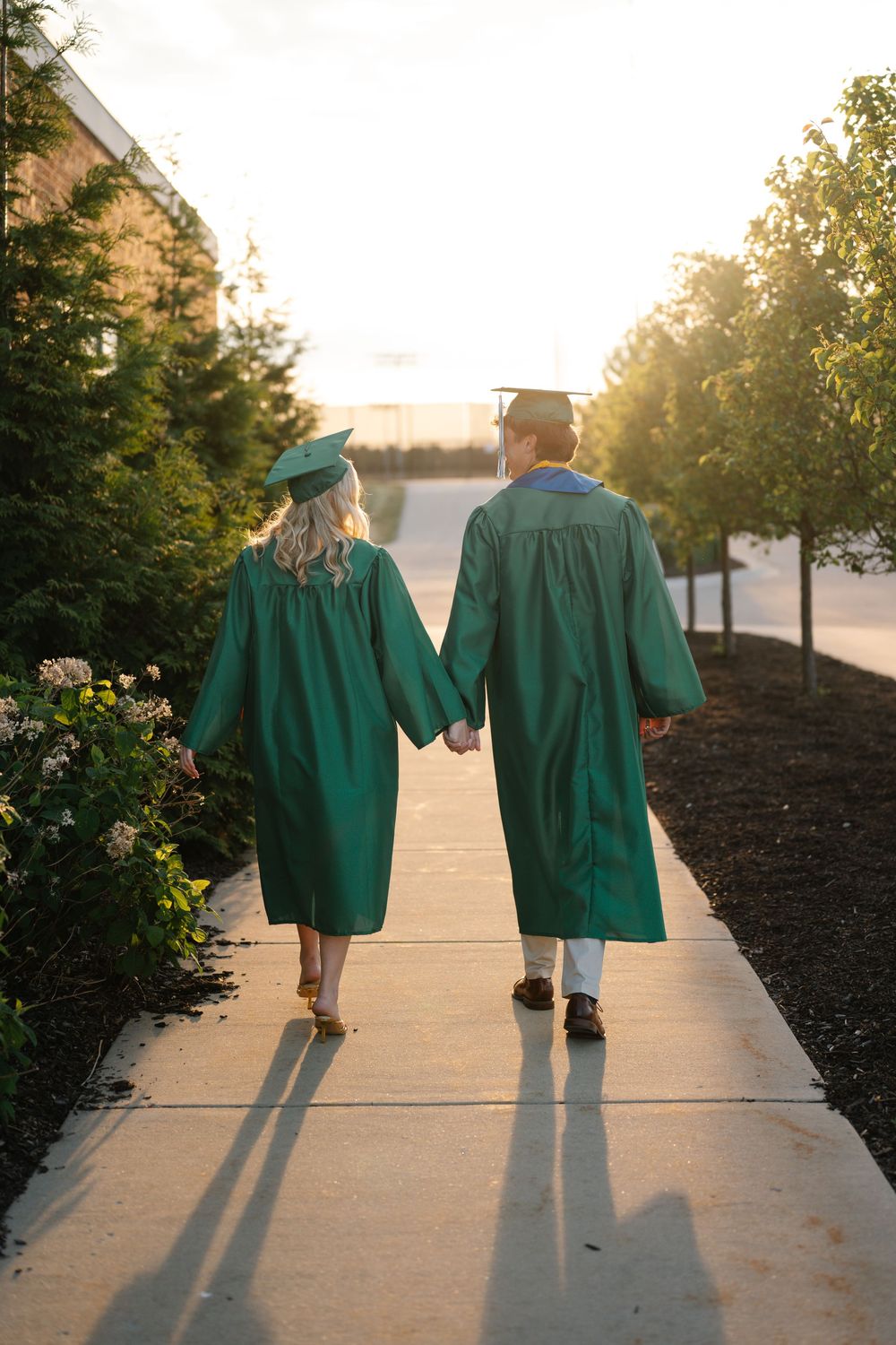 Two graduates in green caps and gowns walking hand in hand down a sunlit pathway.