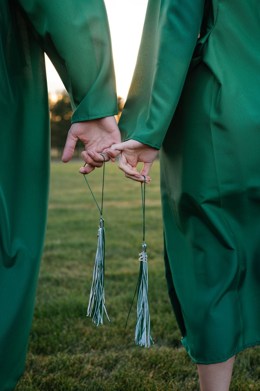 Close-up of two graduates in green gowns holding hands with graduation tassels dangling.