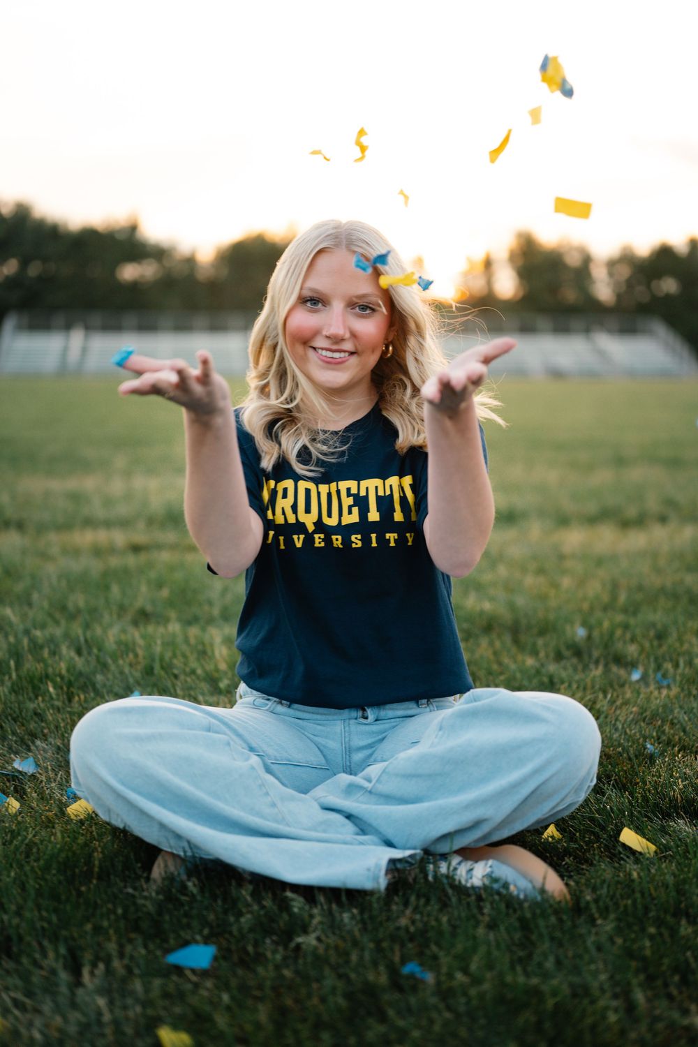 Smiling blonde girl in Marquette University shirt tossing blue and gold confetti on grass.