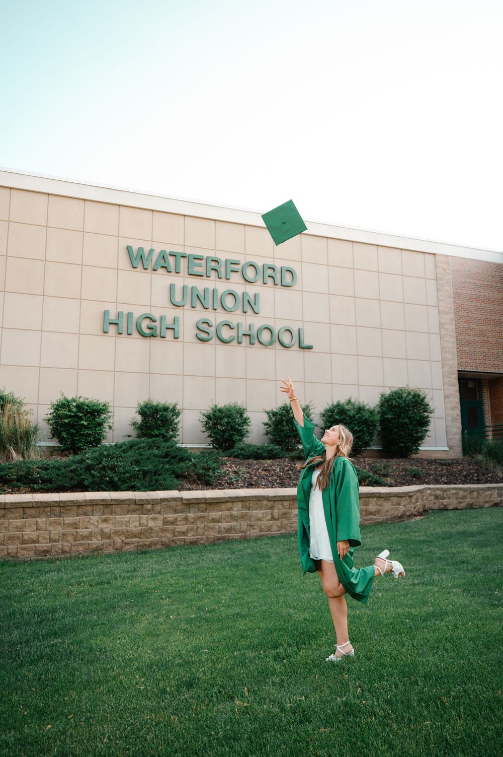 Graduate in green cap and gown tossing cap in front of Waterford Union High School.