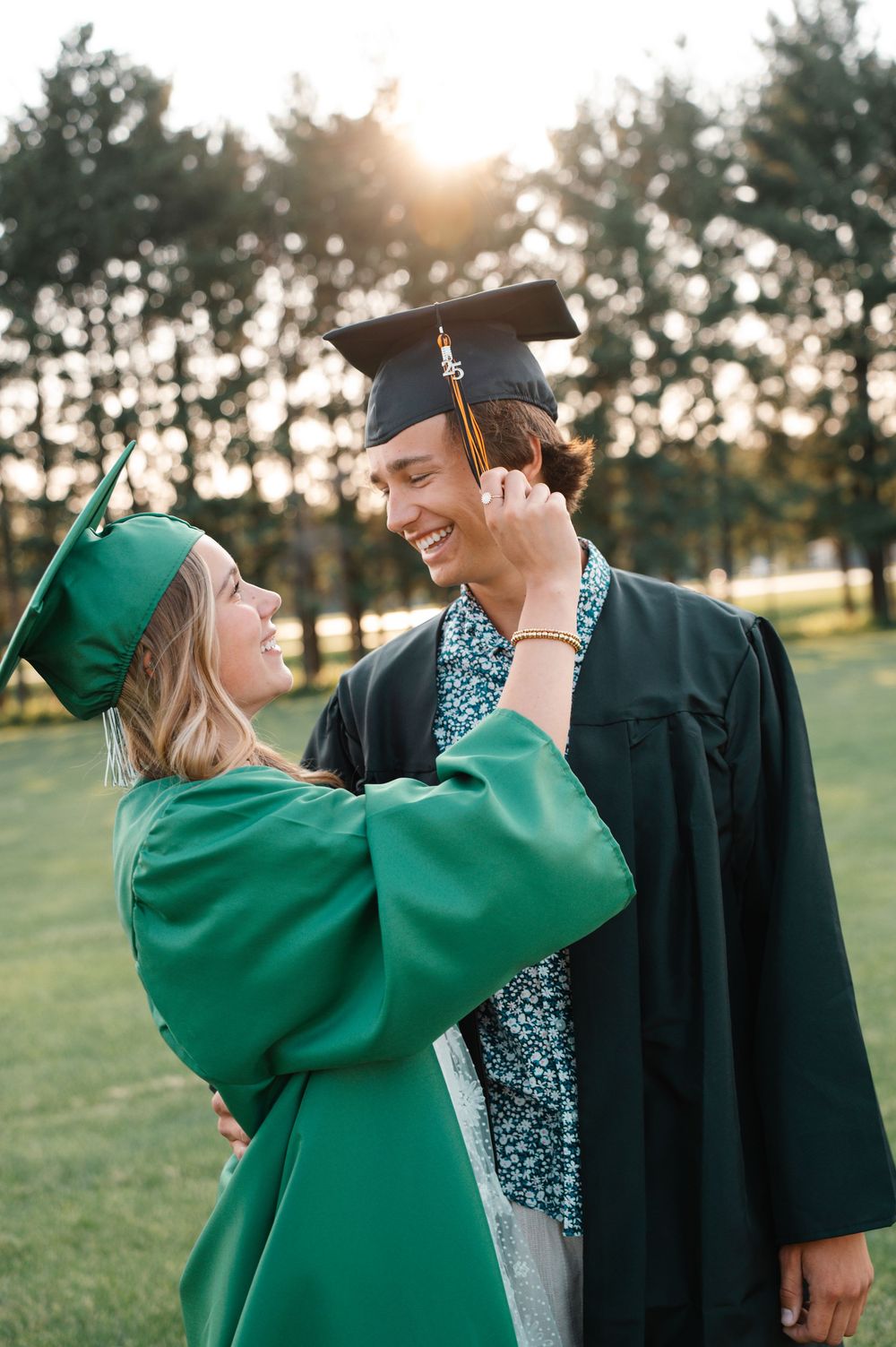 Two graduates in green and black caps and gowns smiling together at golden hour outdoors.
