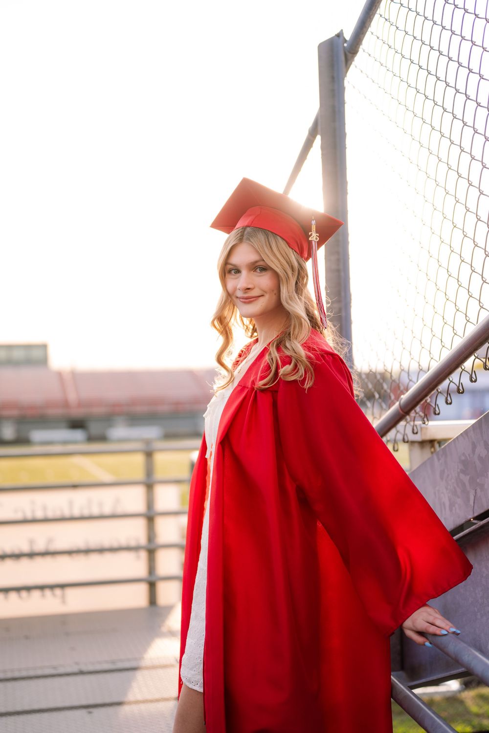 A young woman in red graduation cap and gown poses on stadium bleachers at golden hour.