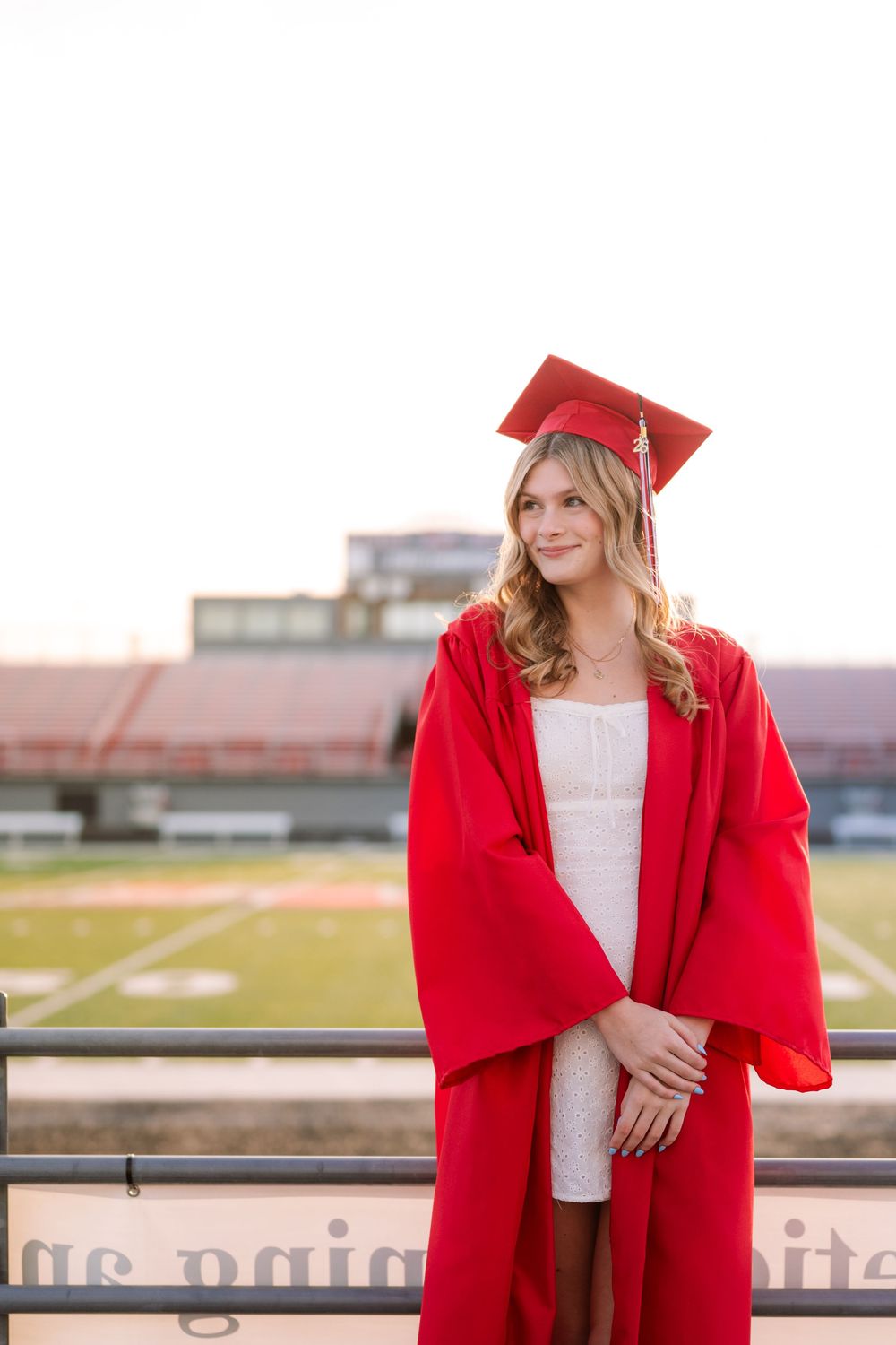 Young woman in red graduation cap and gown posing at a football stadium during golden hour.