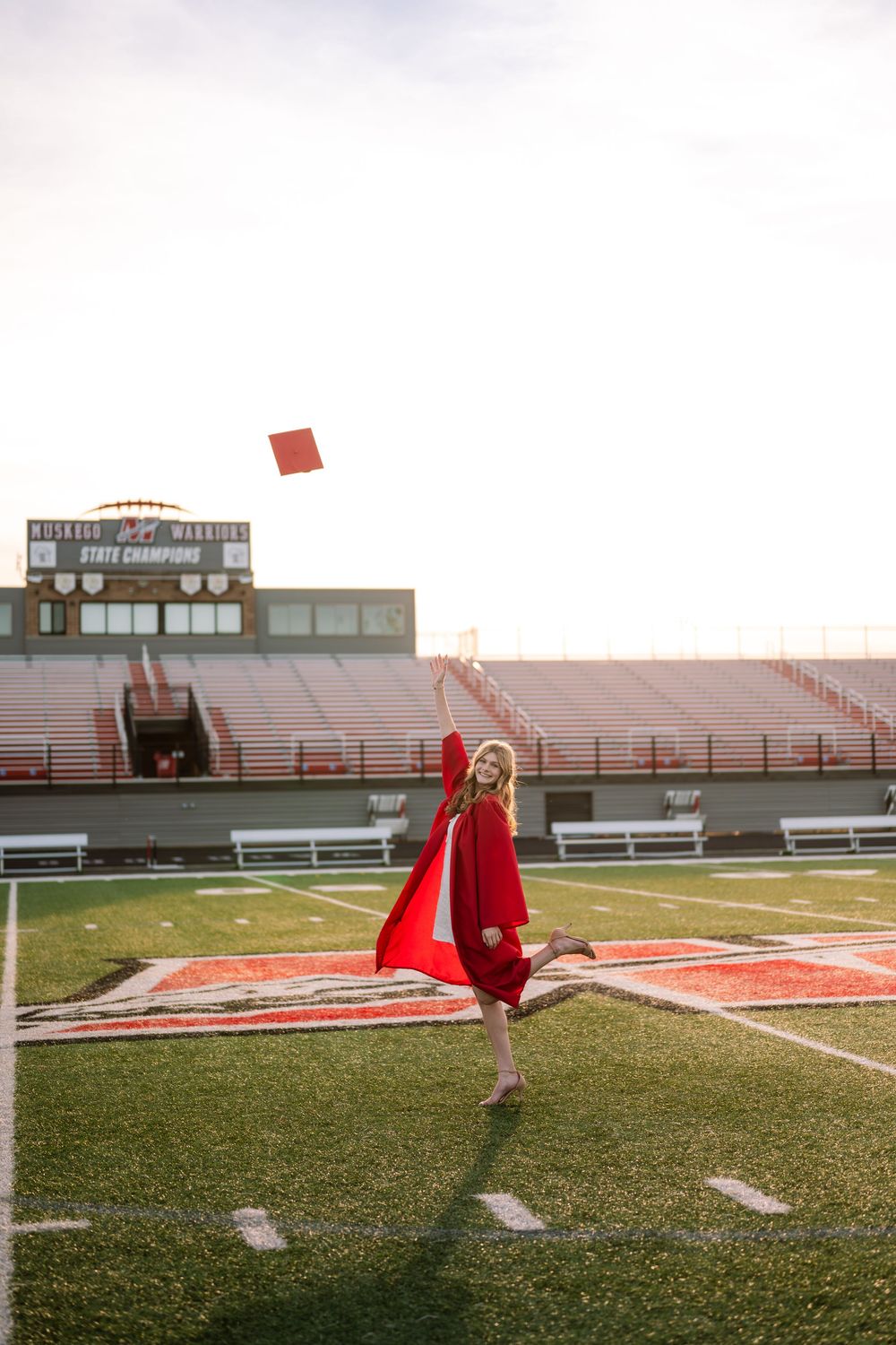 Joyful graduate in red cap and gown tossing her cap on a football field at sunset.