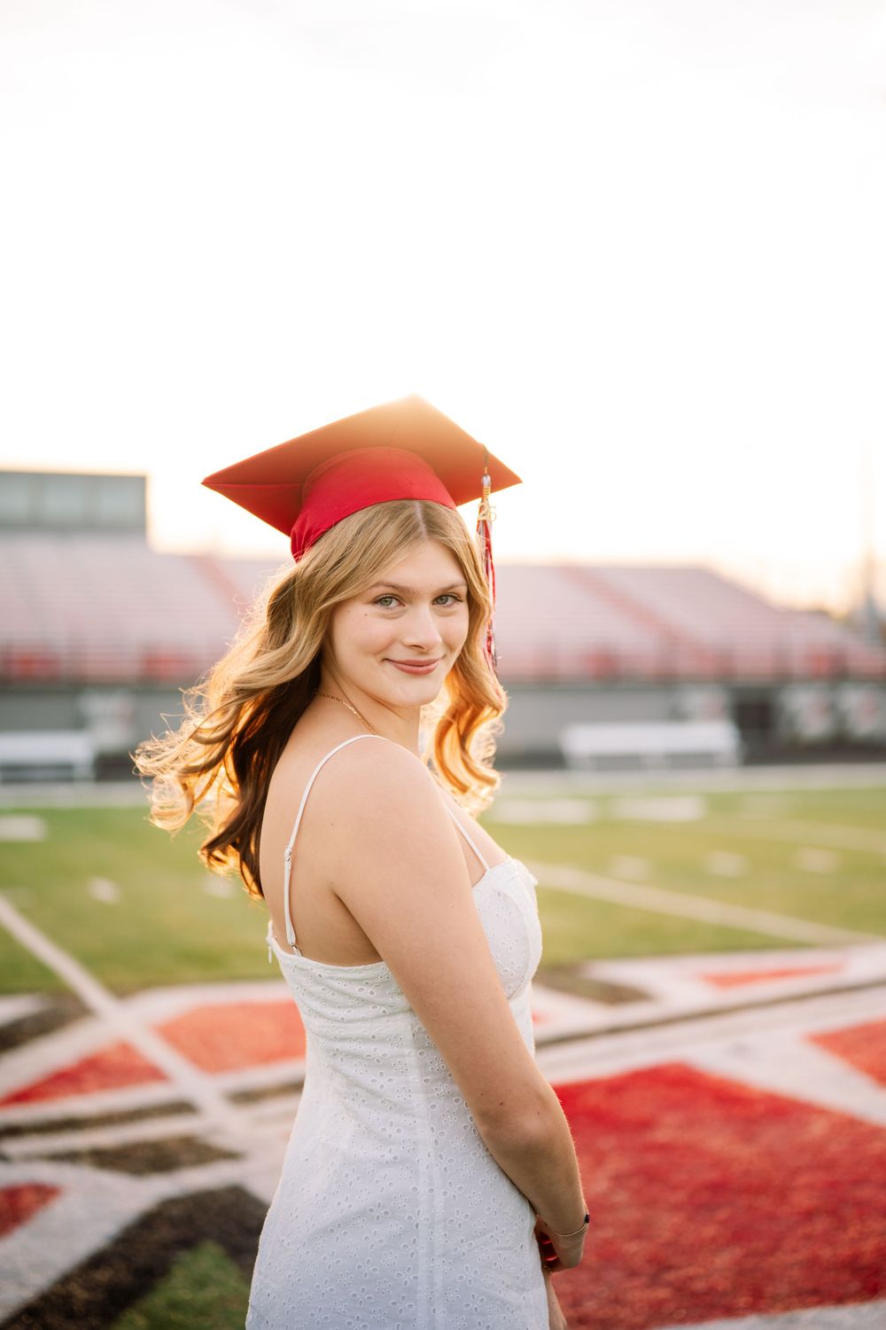 Smiling blonde graduate in red cap and white dress posing on a sunlit football field.