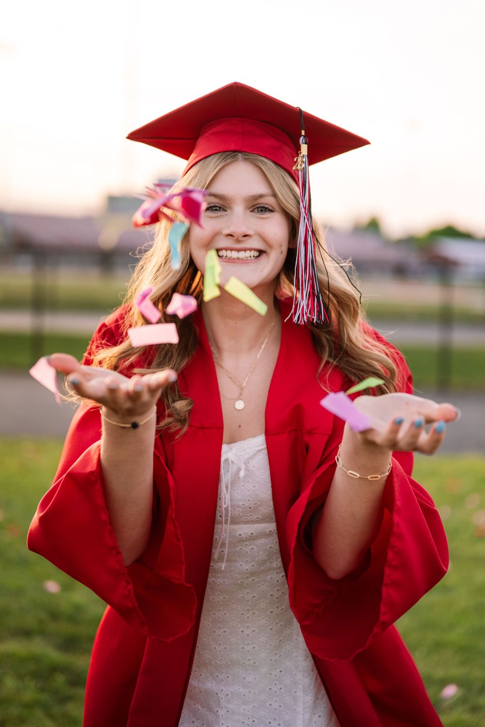 Happy graduate in red cap and gown tossing colorful confetti outdoors at sunset.