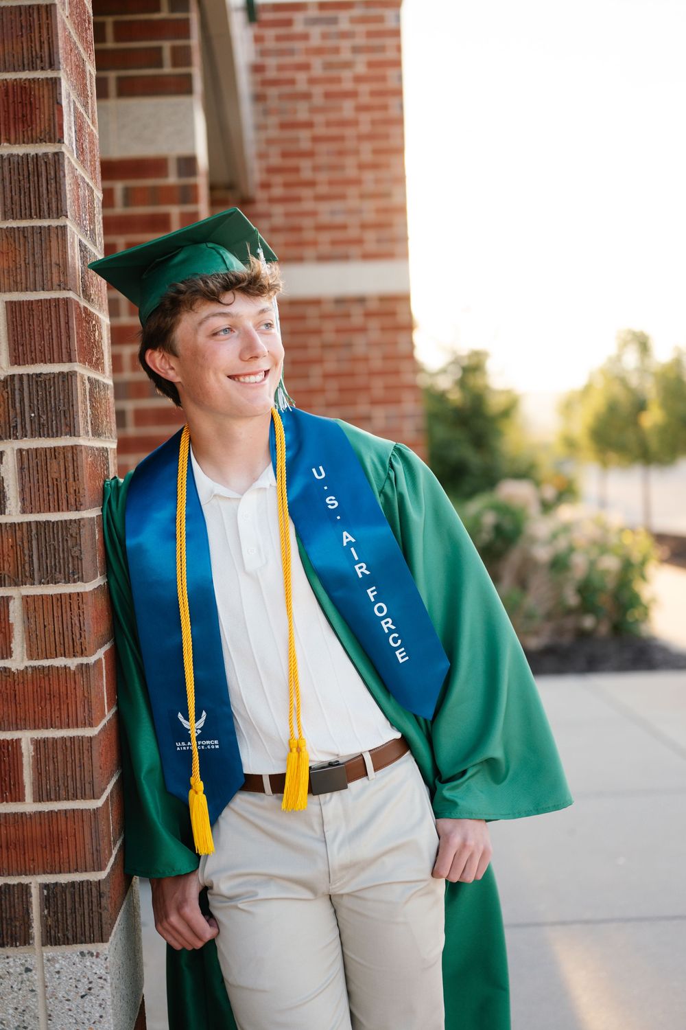 Smiling young man in green graduation gown with U.S. Air Force sash leaning against brick wall.