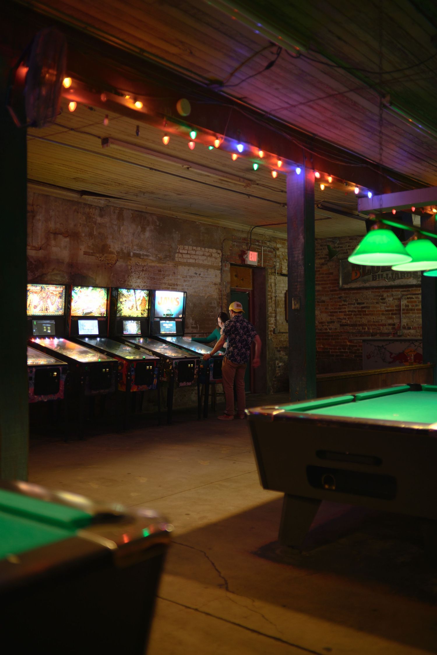 Dark pool hall interior with green felt tables illuminated by colorful string lights and neon signs along brick walls.