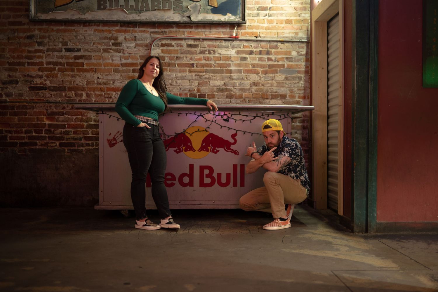Two people posing next to a Red Bull branded wall in an industrial-style space.