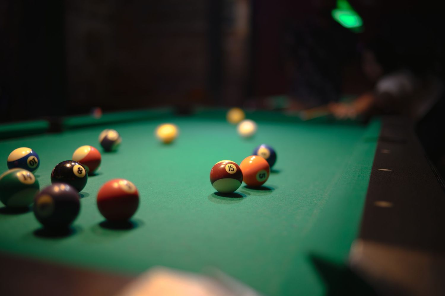 Close-up view of pool balls scattered across a green felt billiards table under moody lighting.