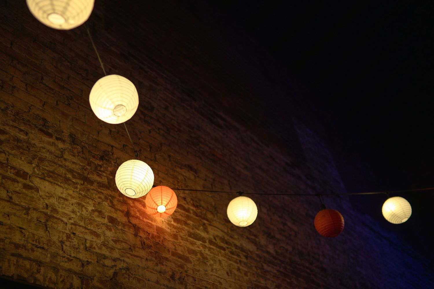 String of glowing paper lanterns illuminated against a brick wall at night.