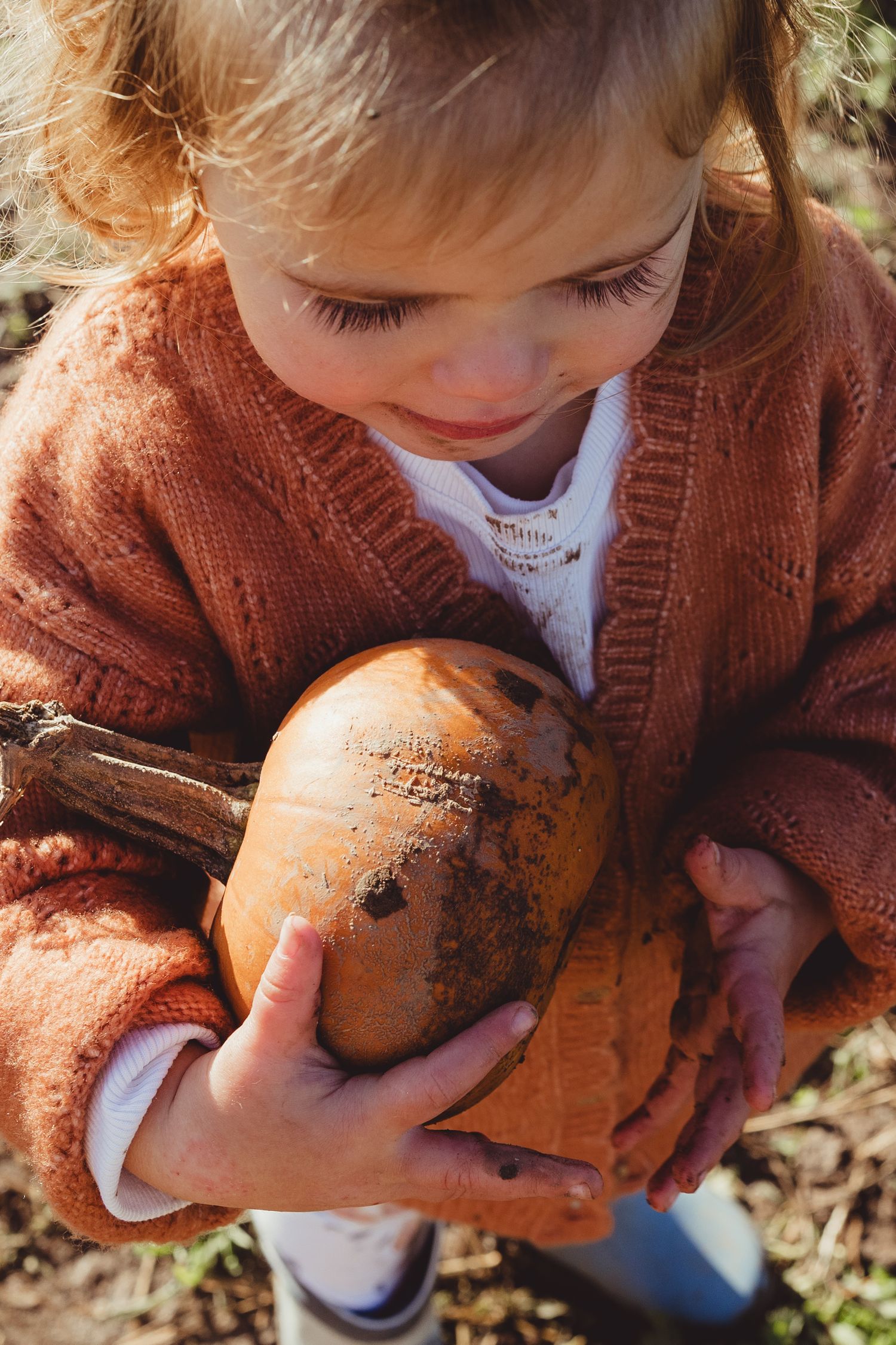 A child in a brown cardigan sweater examines a large dirty sweet potato or root vegetable in an outdoor setting.