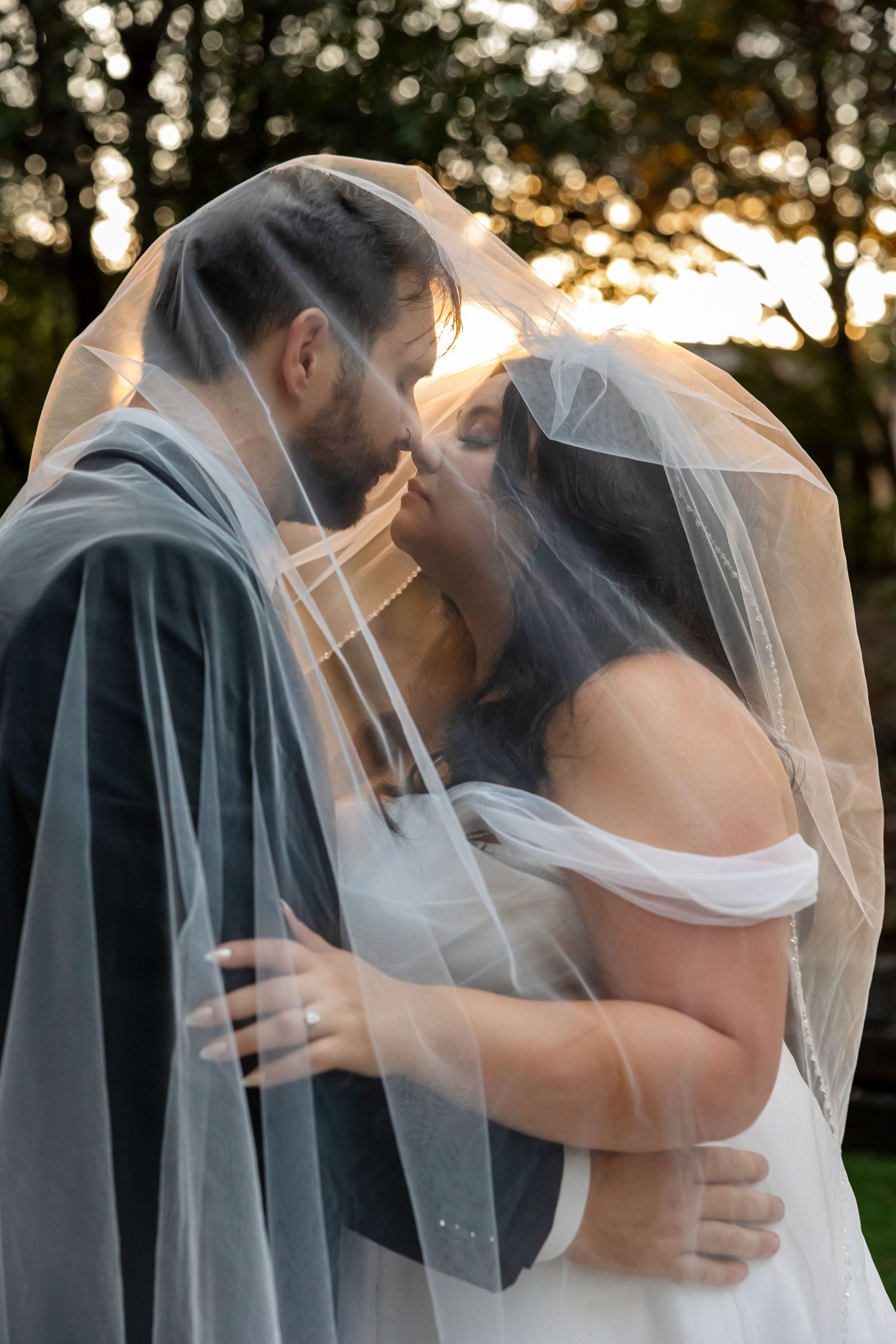 A romantic wedding couple shares an intimate moment under a flowing veil during golden hour sunset.