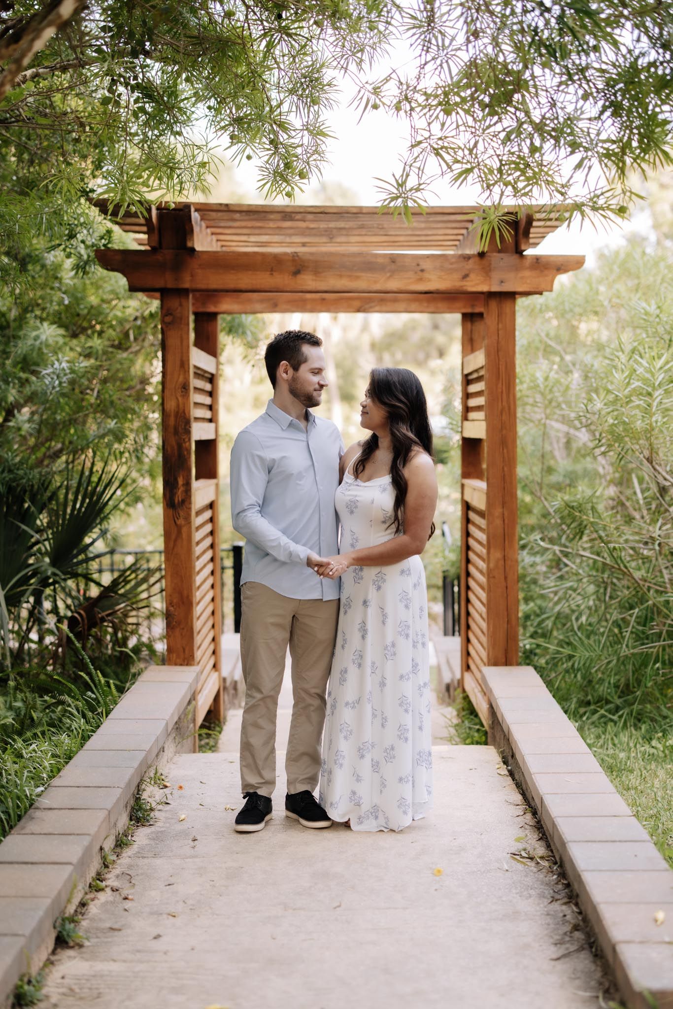A couple stands together under a wooden pergola surrounded by lush greenery in a romantic garden setting.