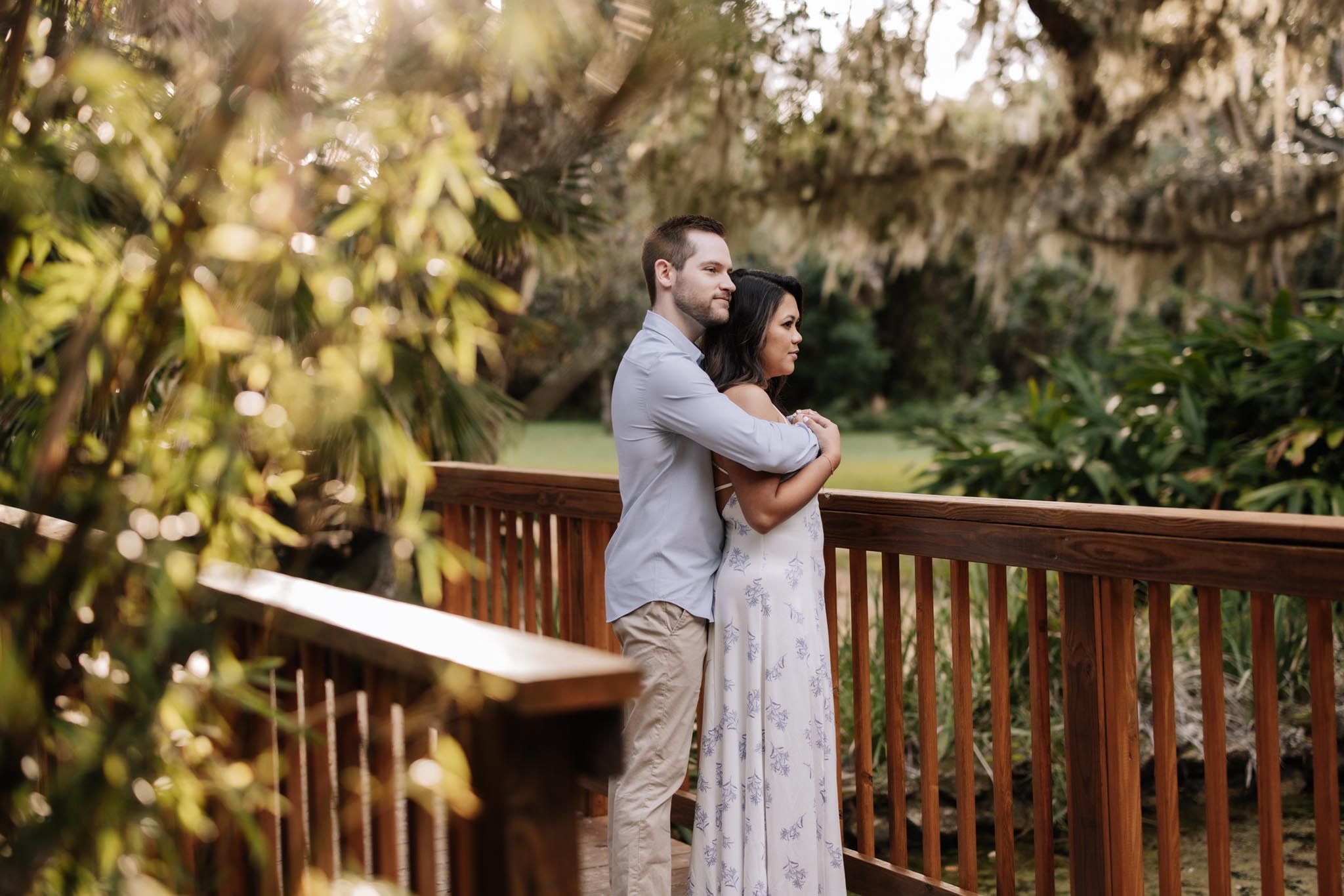 A romantic couple embraces in a lush garden setting with golden sunlight filtering through trees and wooden railings.