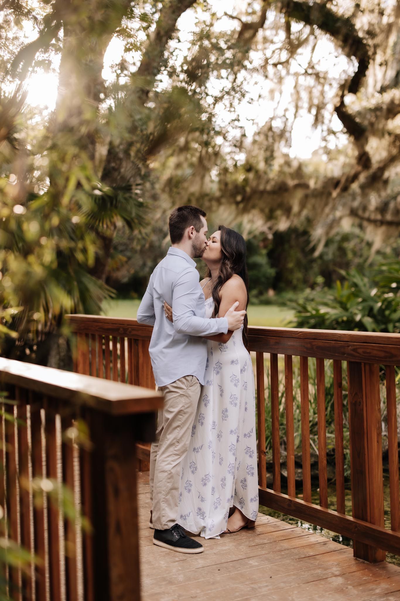 A romantic couple shares an intimate moment on a wooden deck surrounded by Spanish moss and lush greenery at sunset.