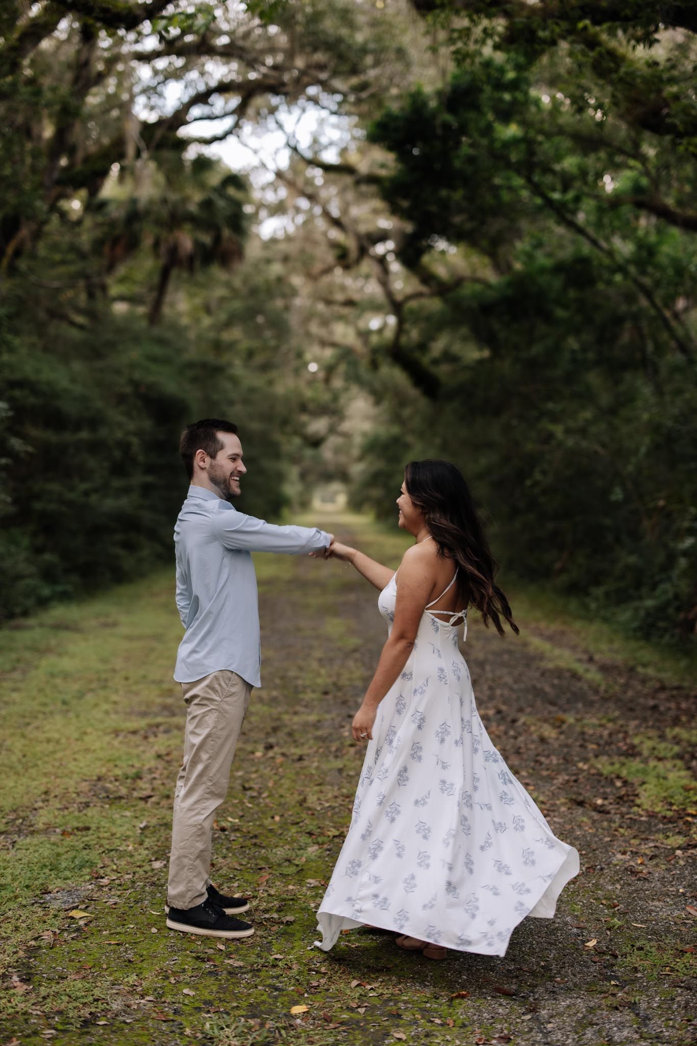 A couple in casual and formal attire dance together on a tree-lined path during an outdoor photo session.