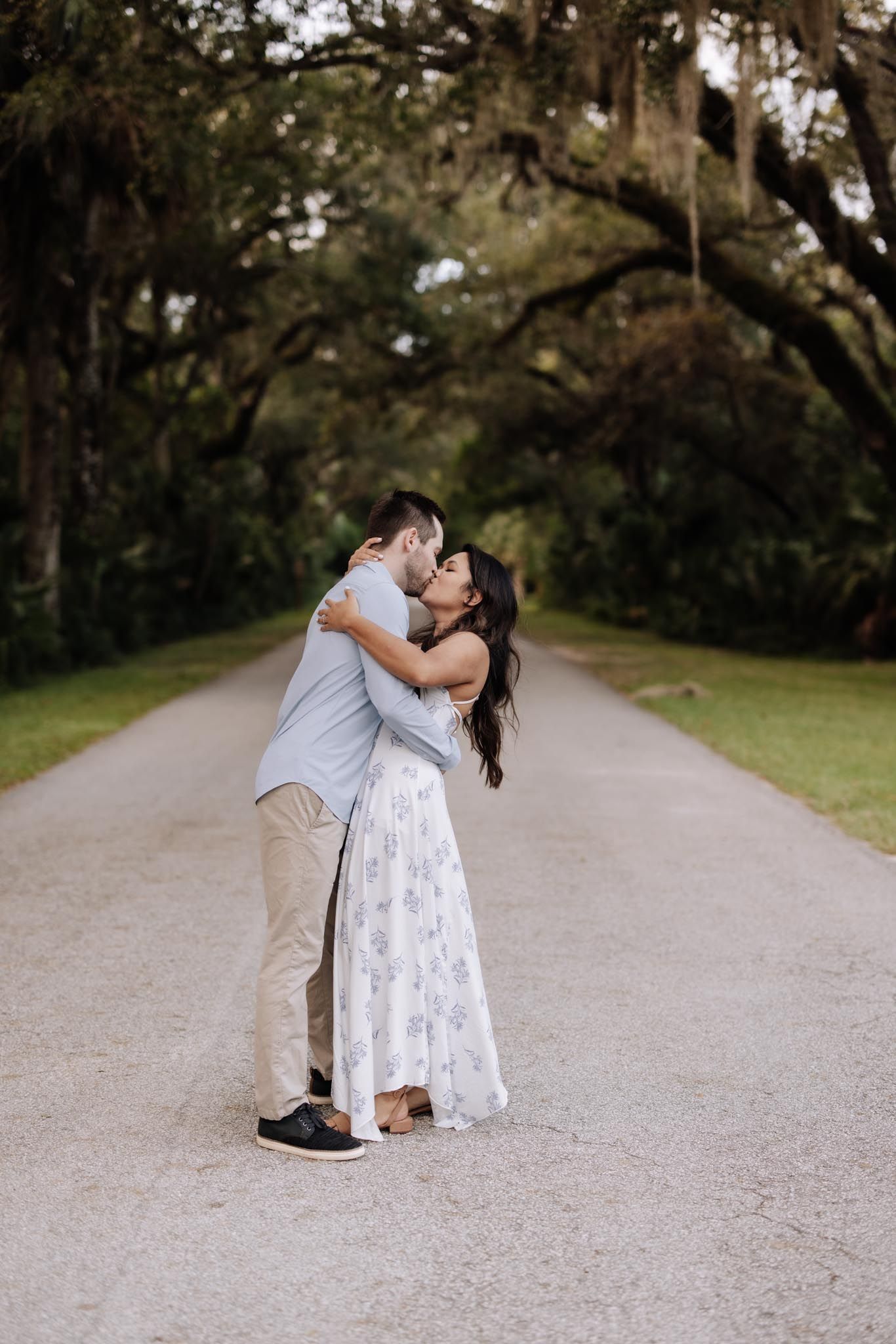 A couple embraces on a tree-lined path with Spanish moss hanging from oak branches overhead.