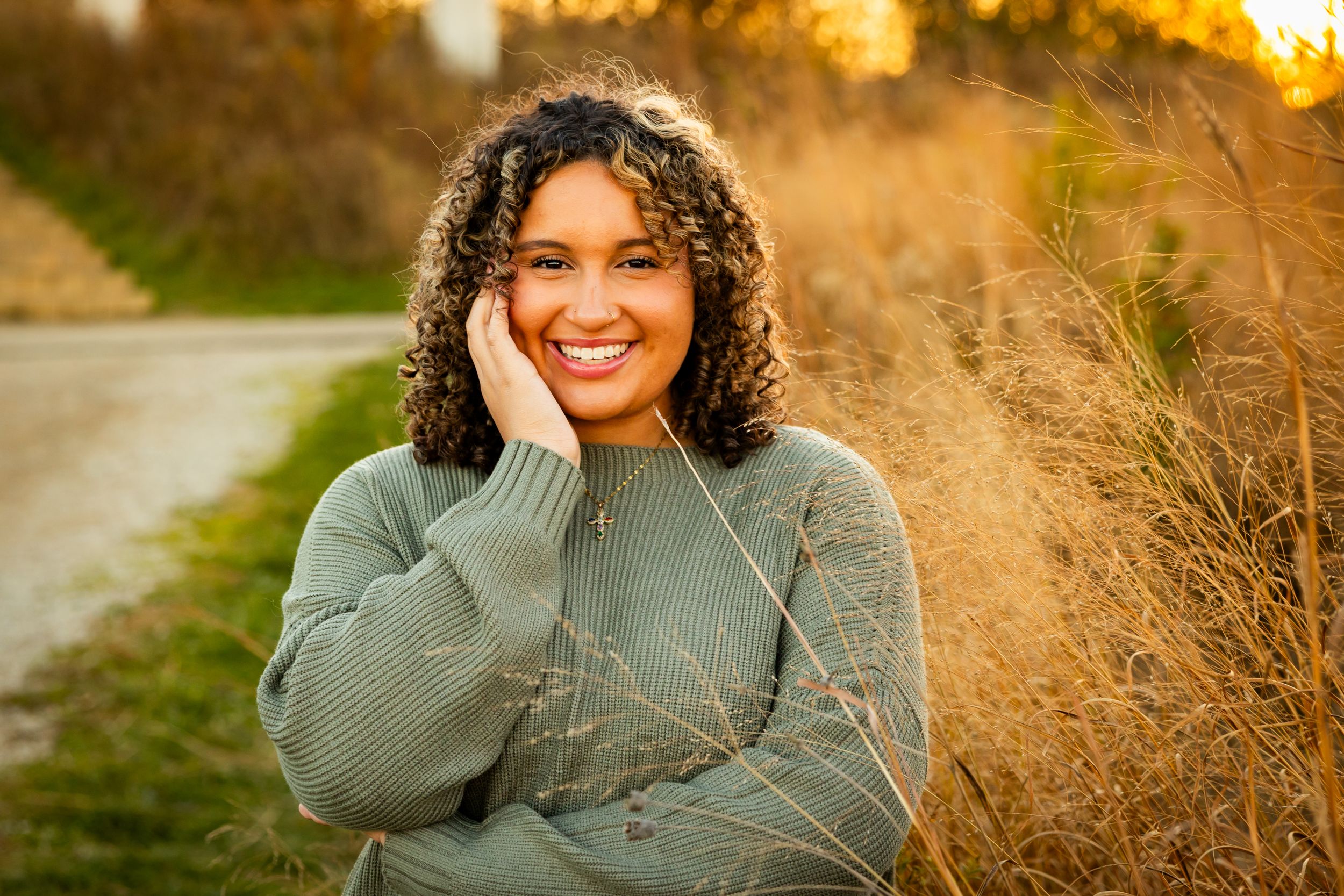Smiling young woman with curly hair in a green sweater posing outdoors among golden grasses at sunset.