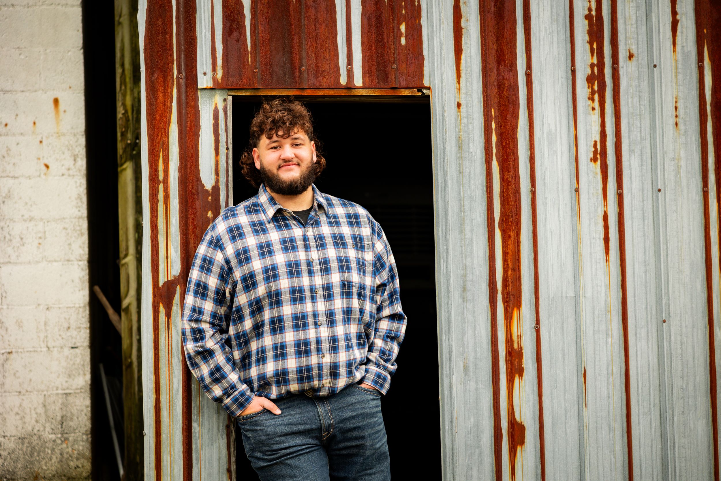 Person in plaid shirt and jeans poses against rustic metal wall with peeling paint and weathered texture.