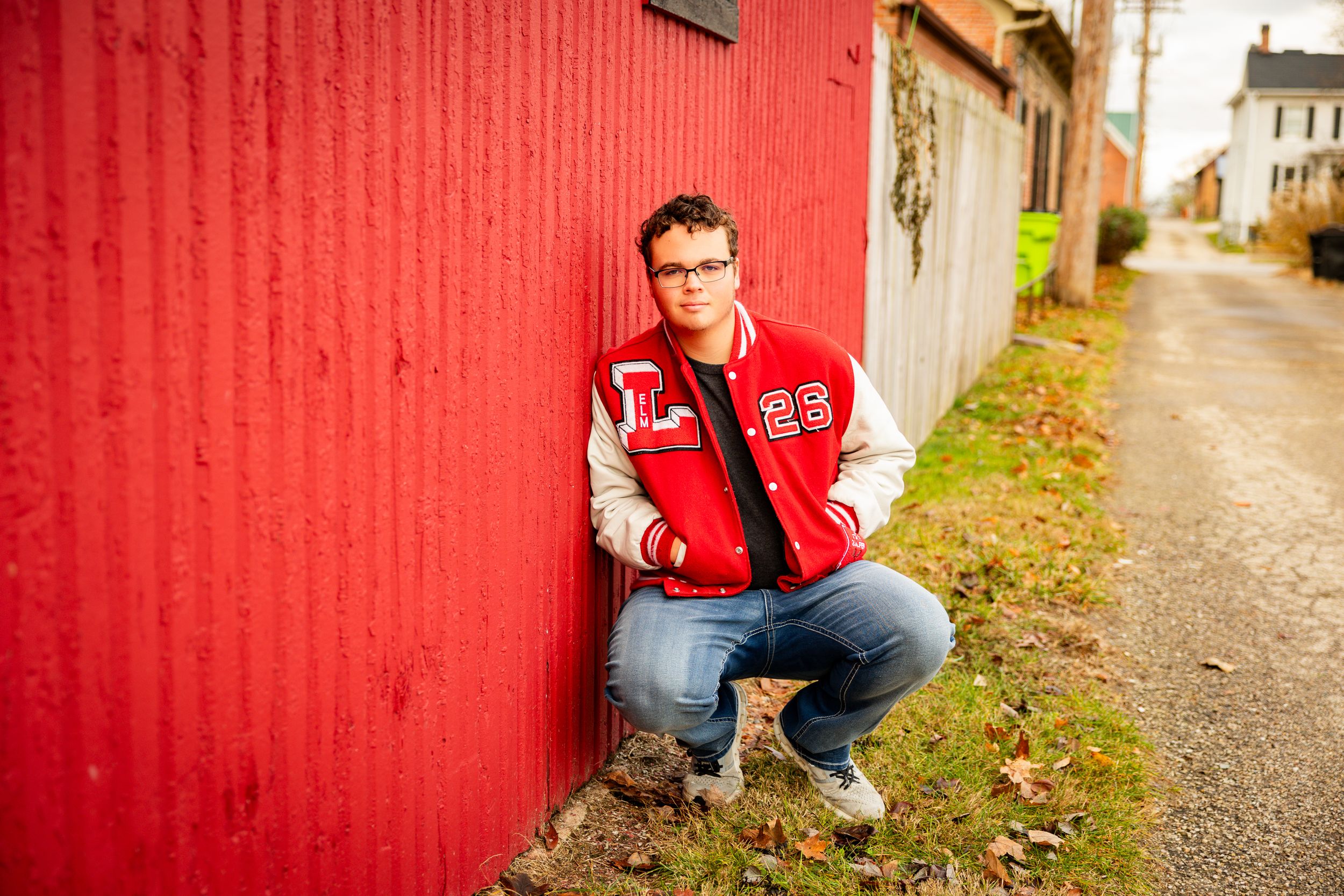 Someone in a red varsity jacket crouches next to a bright red wall along a narrow path.
