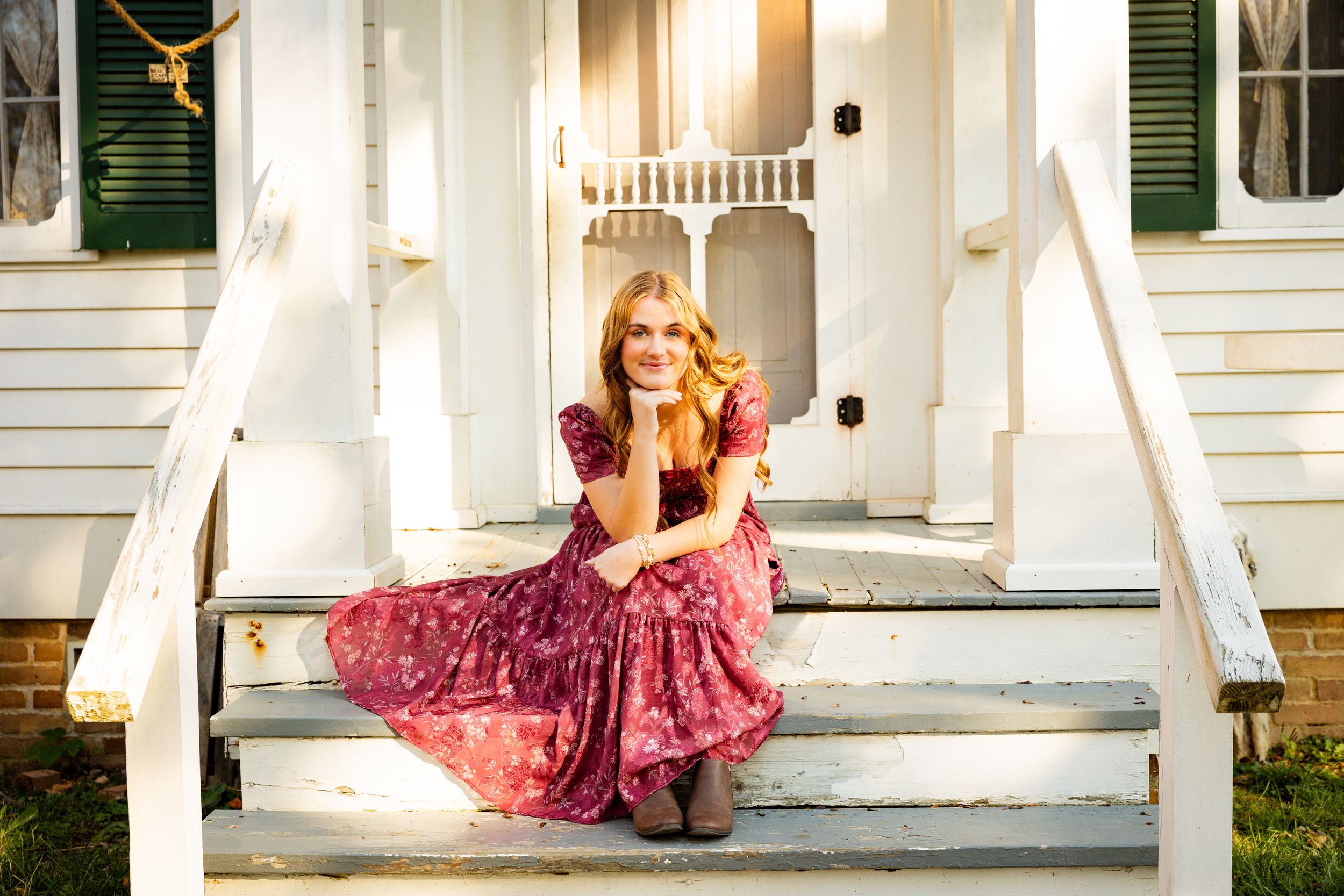 A person in a flowing pink dress poses on the steps of a white house with green shutters.