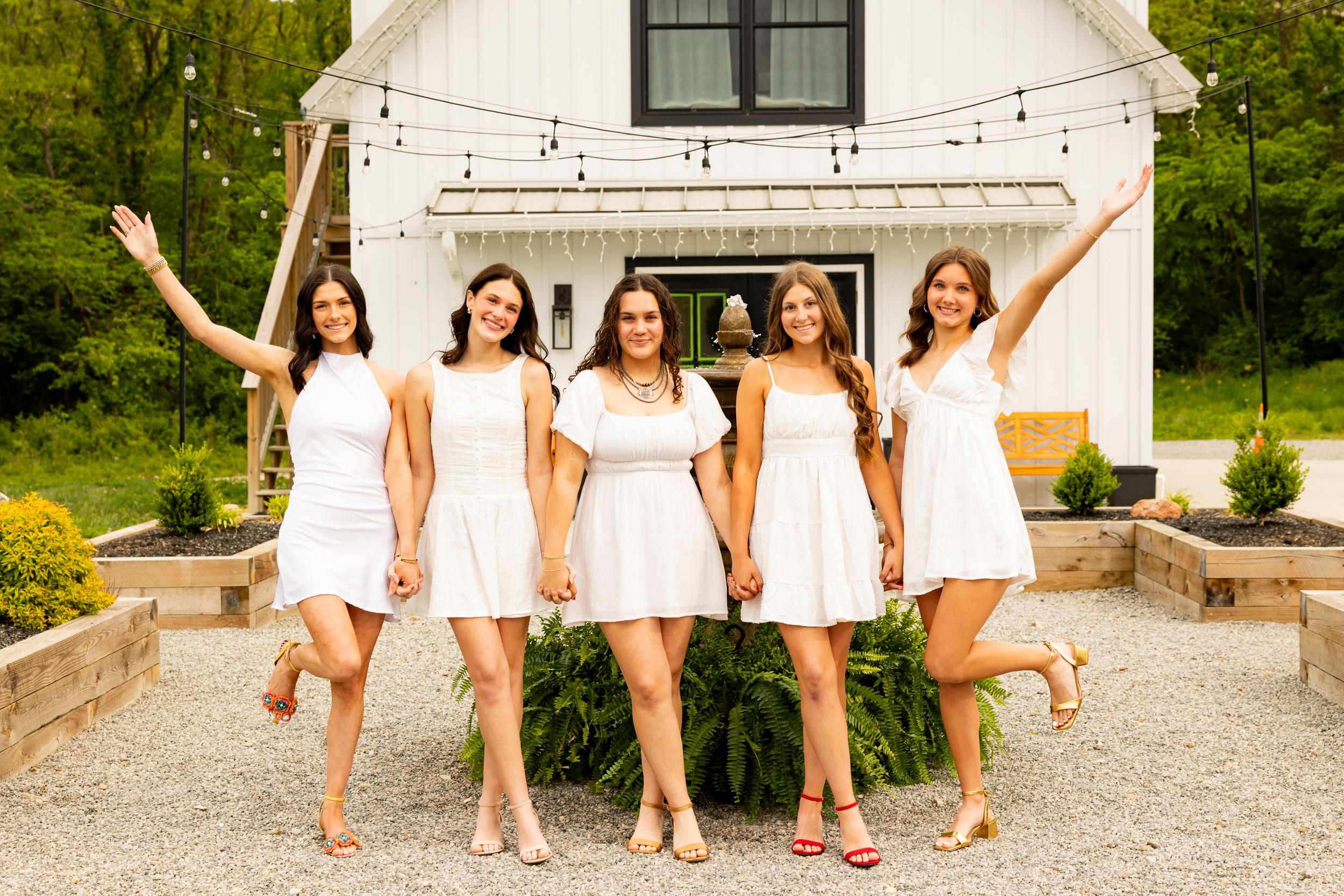Five young women in white dresses pose joyfully outside a white barn venue.