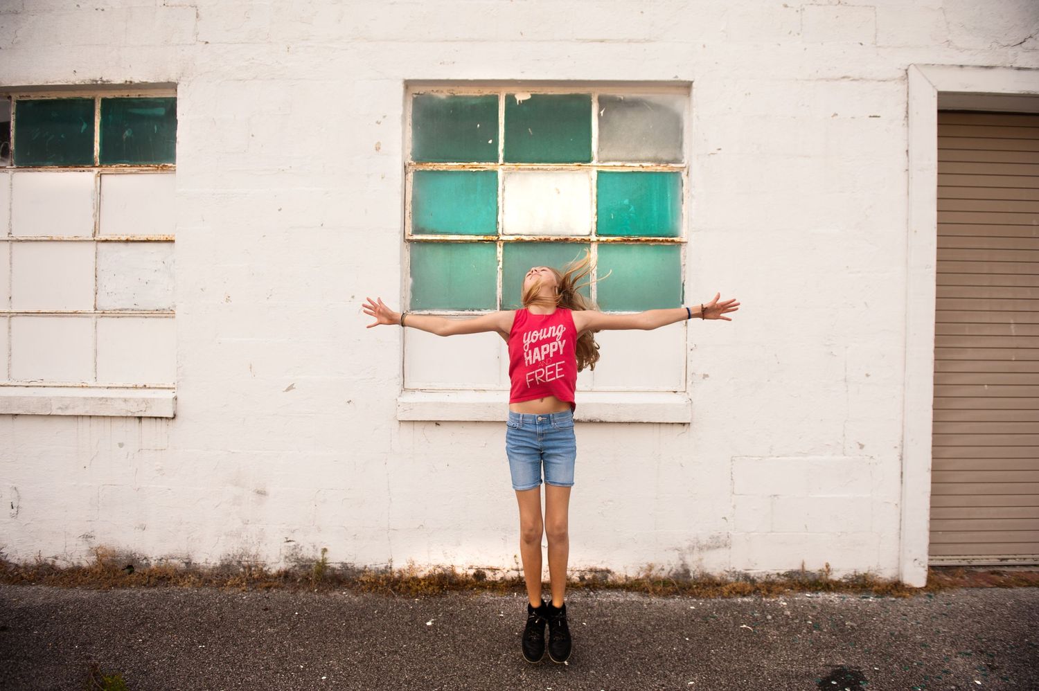 Person in a red shirt and shorts stands with arms outstretched against a white wall with green-trimmed windows.