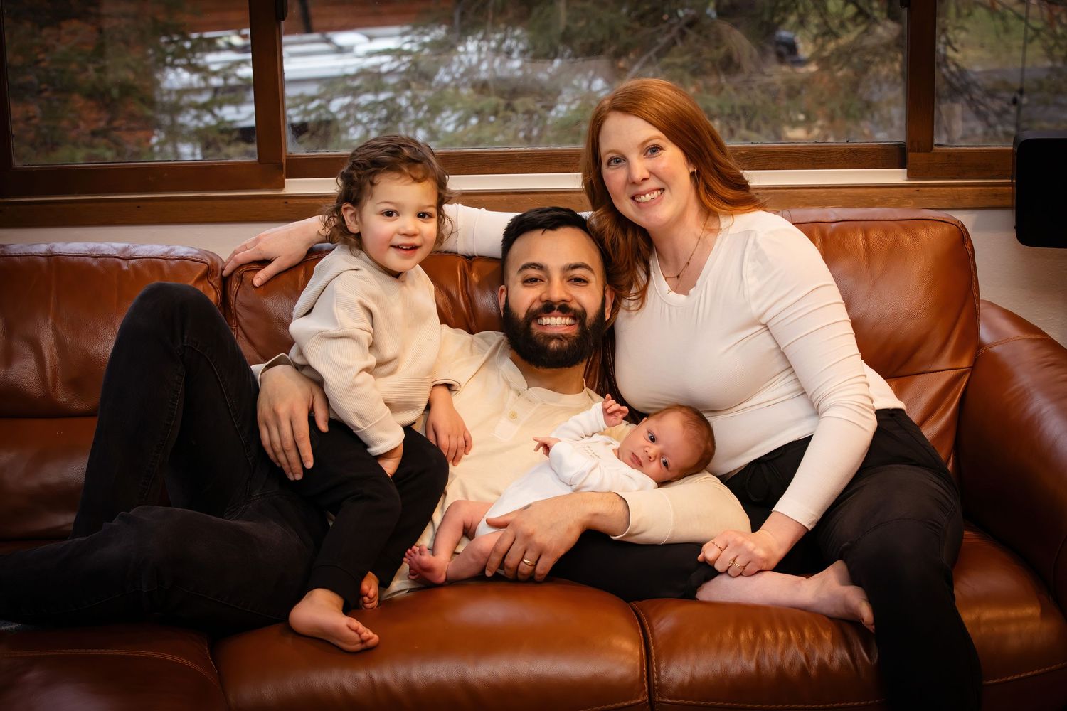 Intimate family portrait on brown leather couch with natural window light.