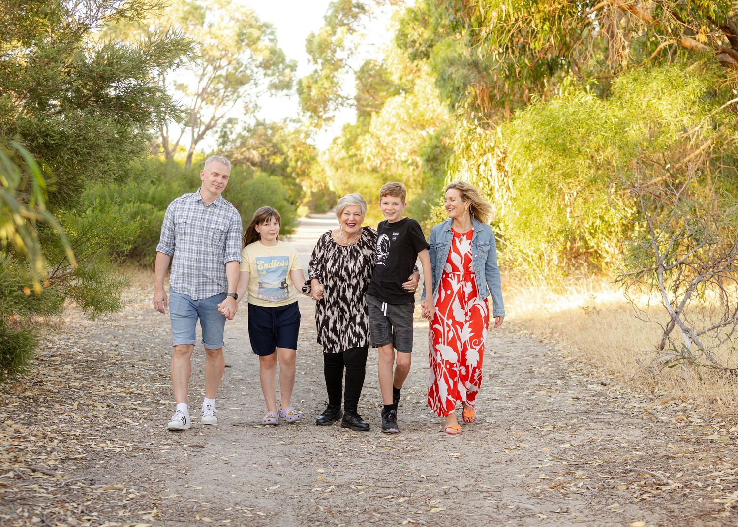 A group of people walking together on a dirt path surrounded by lush green trees on a sunny afternoon.