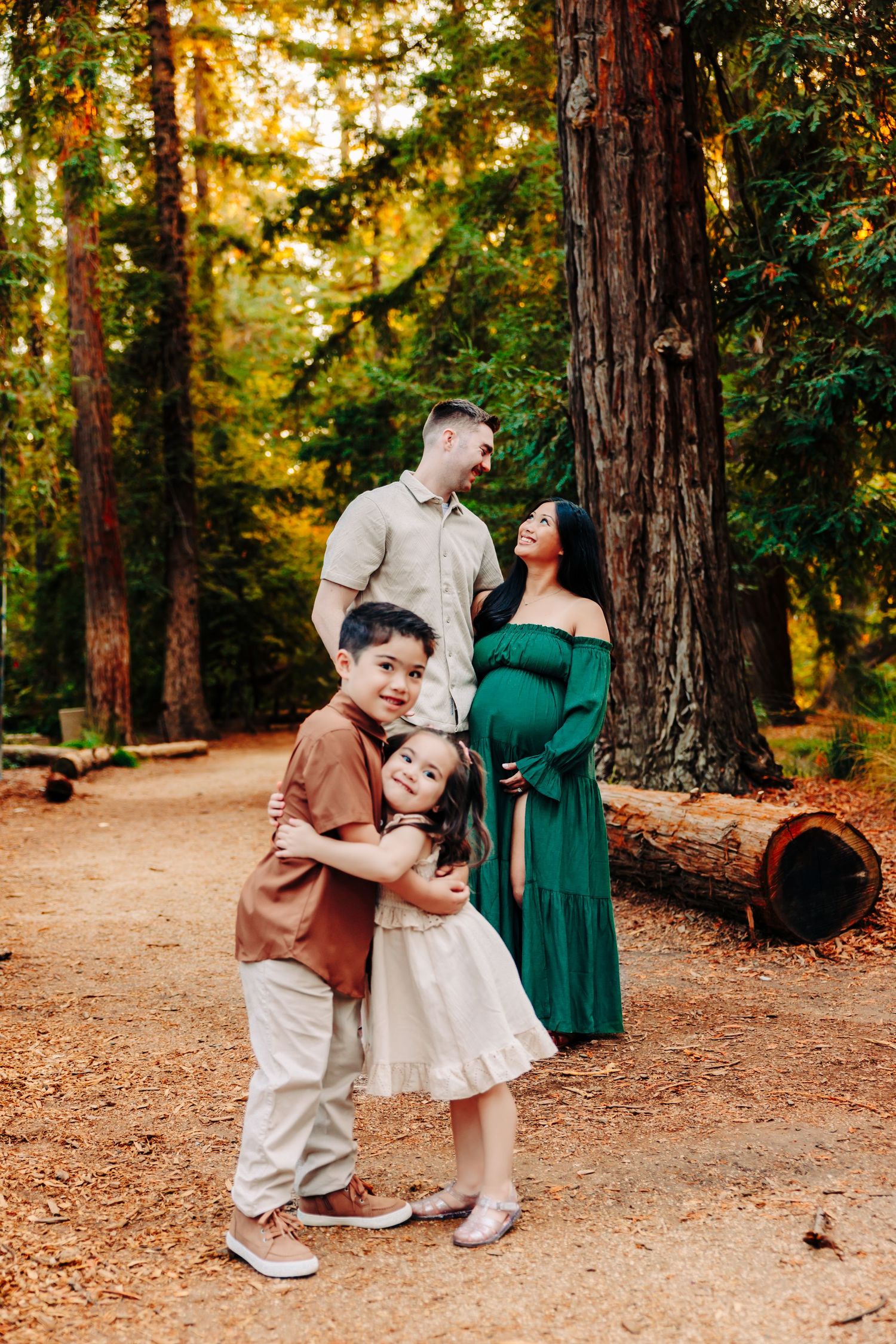 A family enjoys a tender moment together on a forest path surrounded by tall redwood trees at sunset.