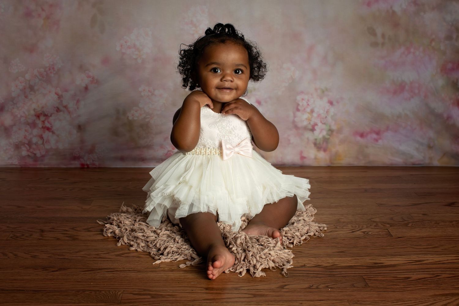 A small child in a white lace dress sits on a wooden floor while posing in different playful positions.