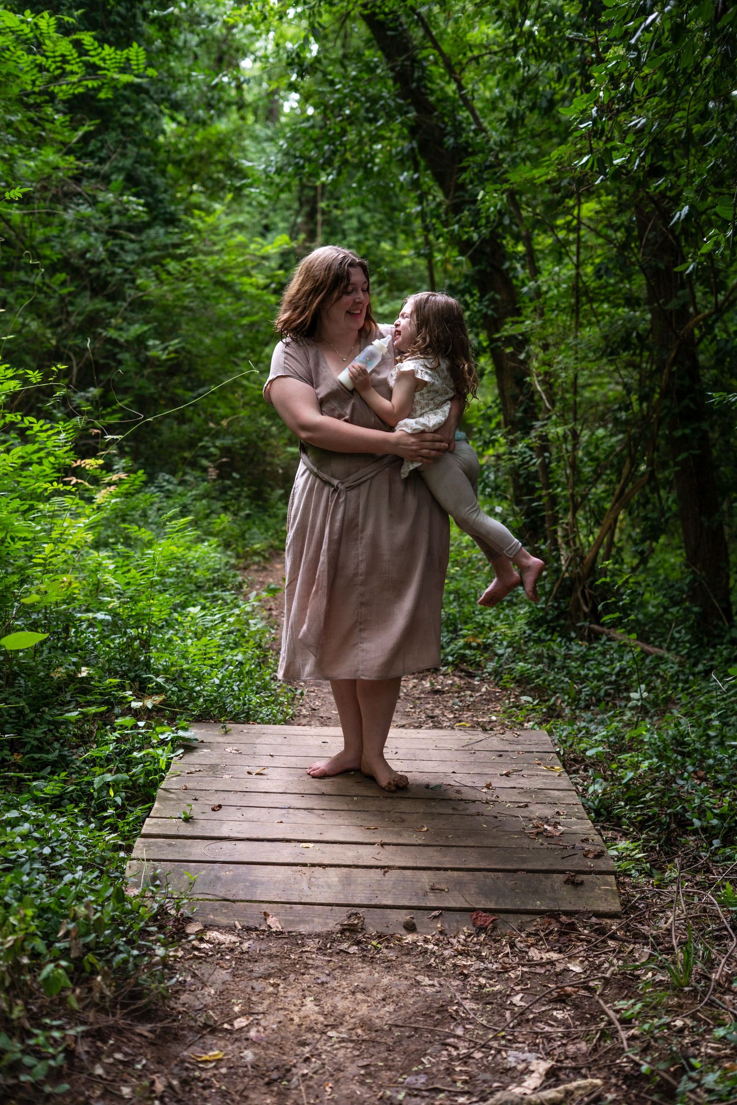 A person in a beige dress sits peacefully on a wooden boardwalk surrounded by lush green forest vegetation.