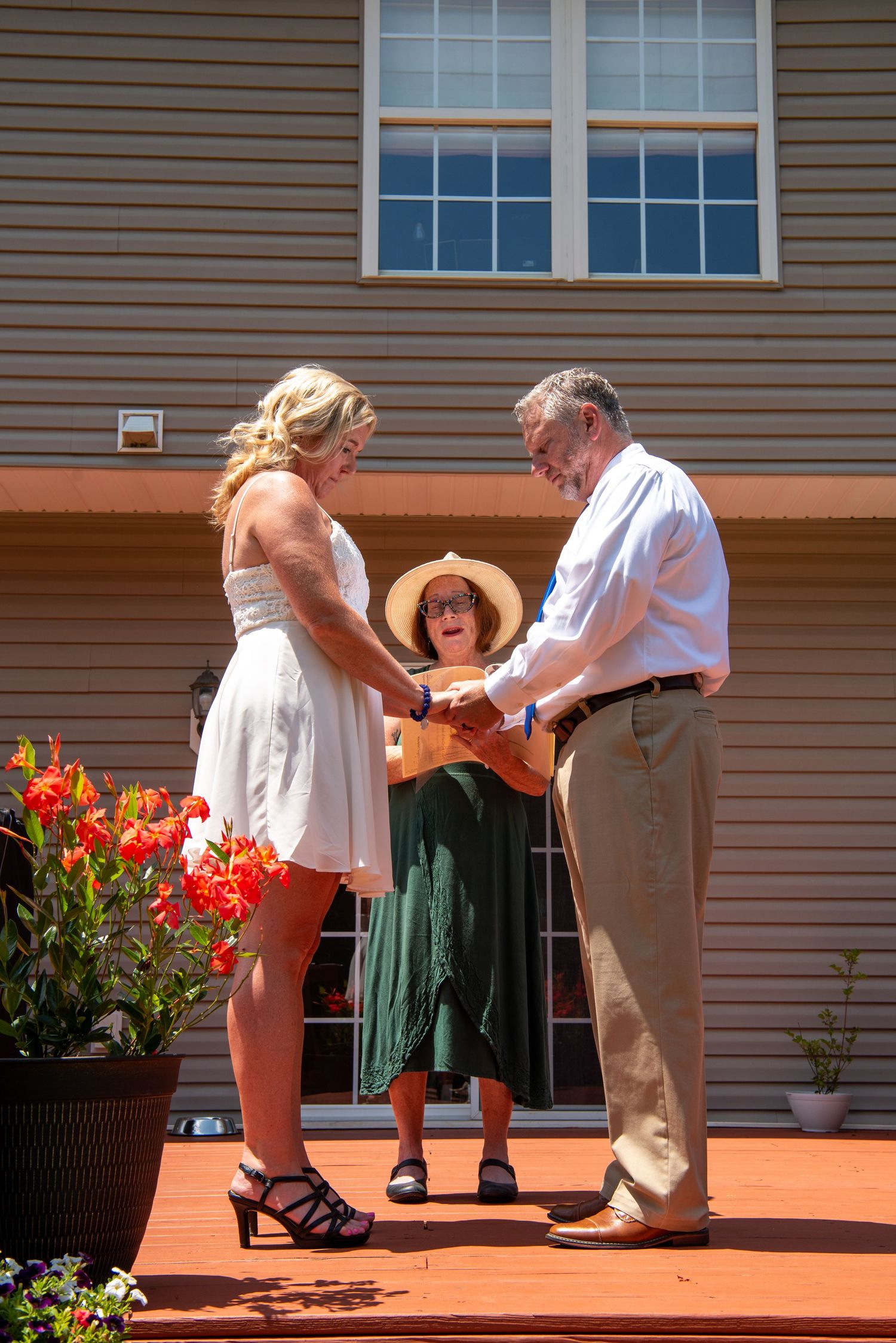 A wedding ceremony unfolds on an orange deck with colorful flowers in planters beside a tan house.