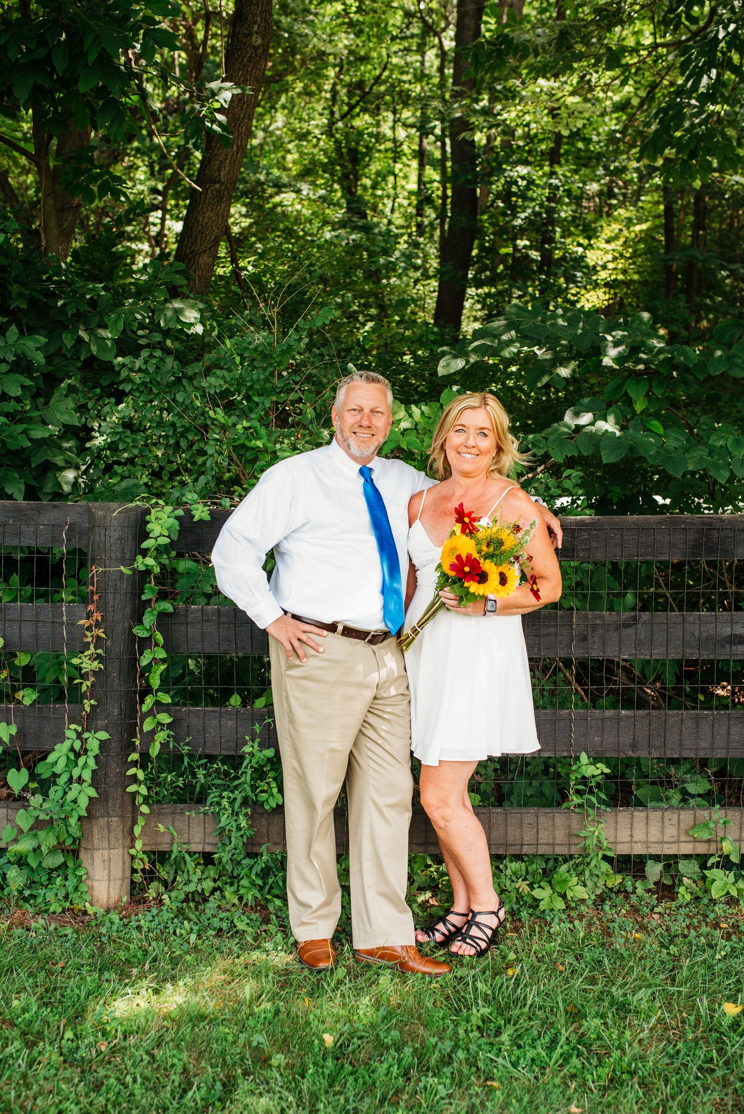 A couple poses together in front of a wooden fence surrounded by lush greenery on a sunny summer day.