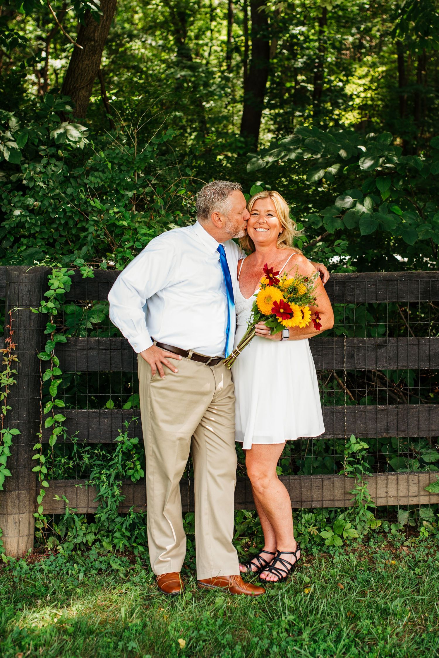 A couple poses playfully in front of a rustic wooden fence surrounded by lush greenery.