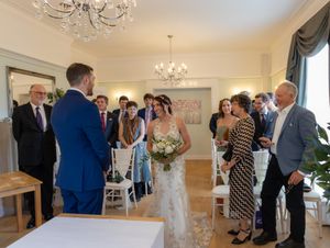 An intimate indoor wedding ceremony takes place in an elegant room with chandeliers and gathered guests.
