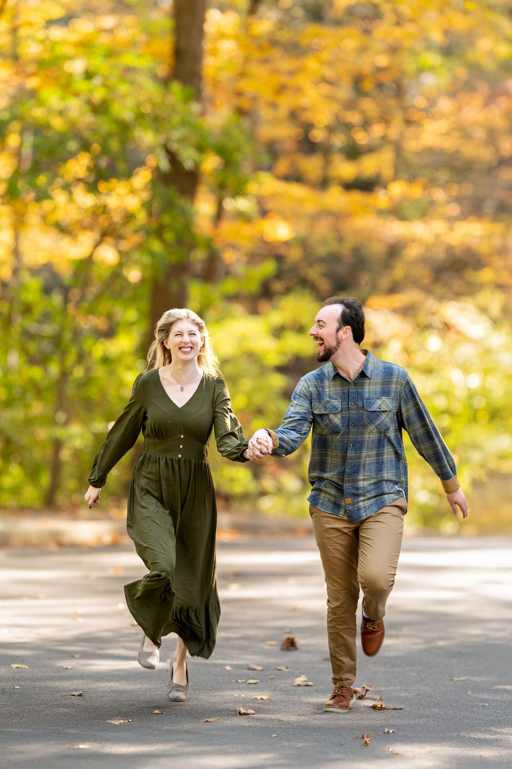 Joyful couple running hand-in-hand toward camera with vibrant yellow fall foliage behind them.