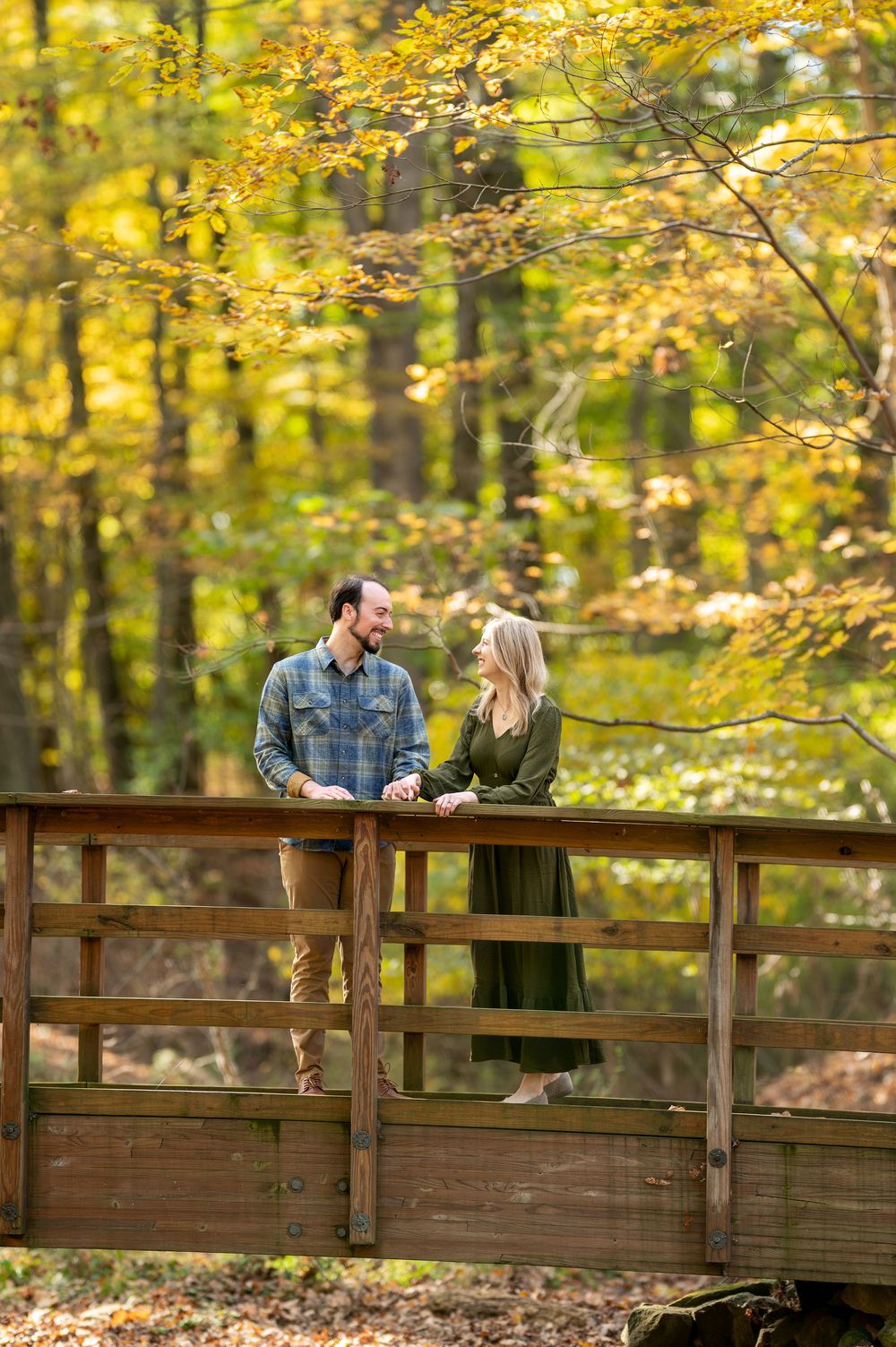 Couple standing on a wooden bridge smiling at each other surrounded by golden autumn trees.