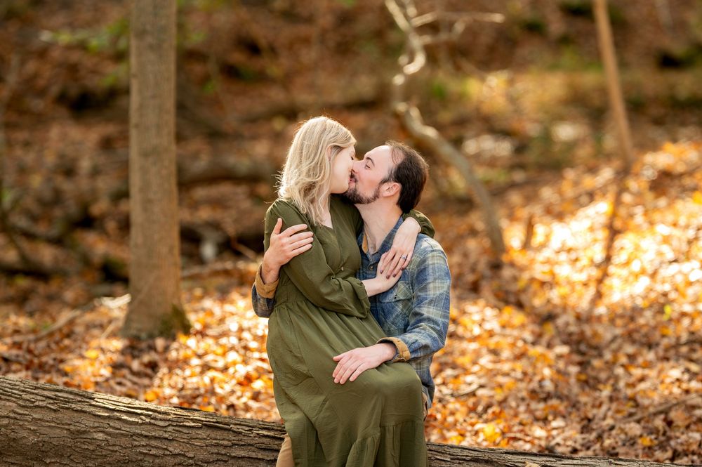 Couple sharing a tender kiss while seated on a fallen log among colorful autumn leaves.