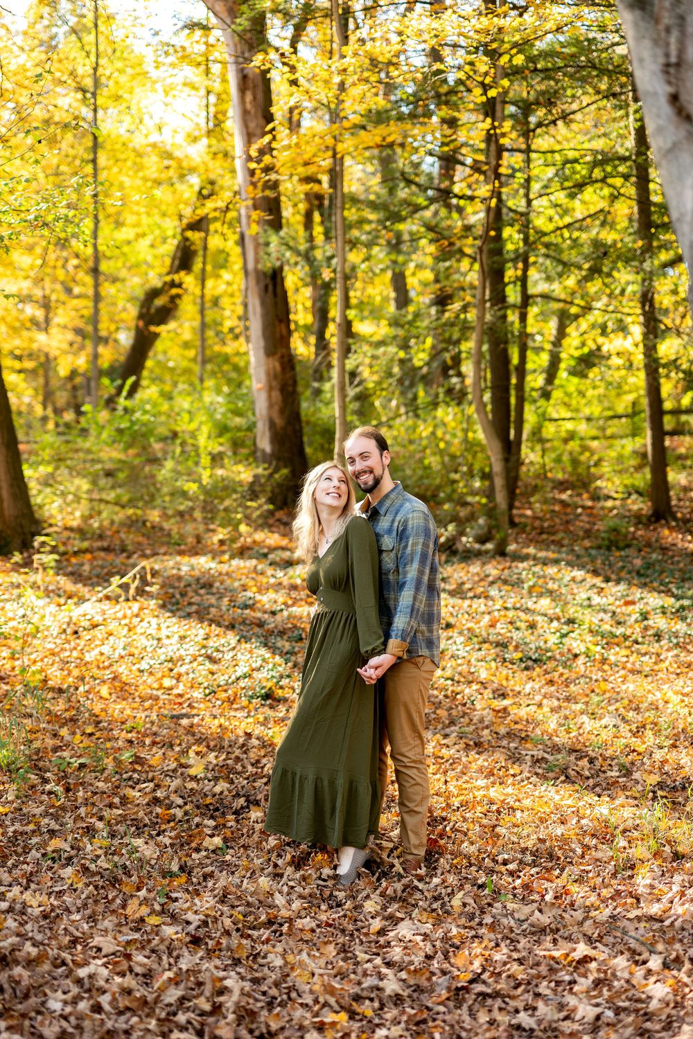 Smiling engaged couple holding hands and posing together in a bright yellow autumn forest.