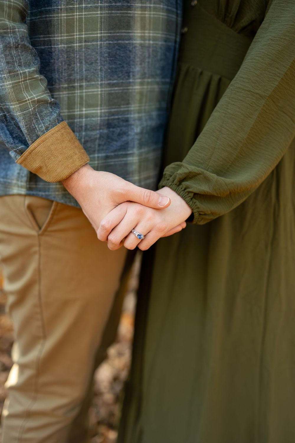 Close-up of engaged couple holding hands showing diamond ring with fall foliage background.